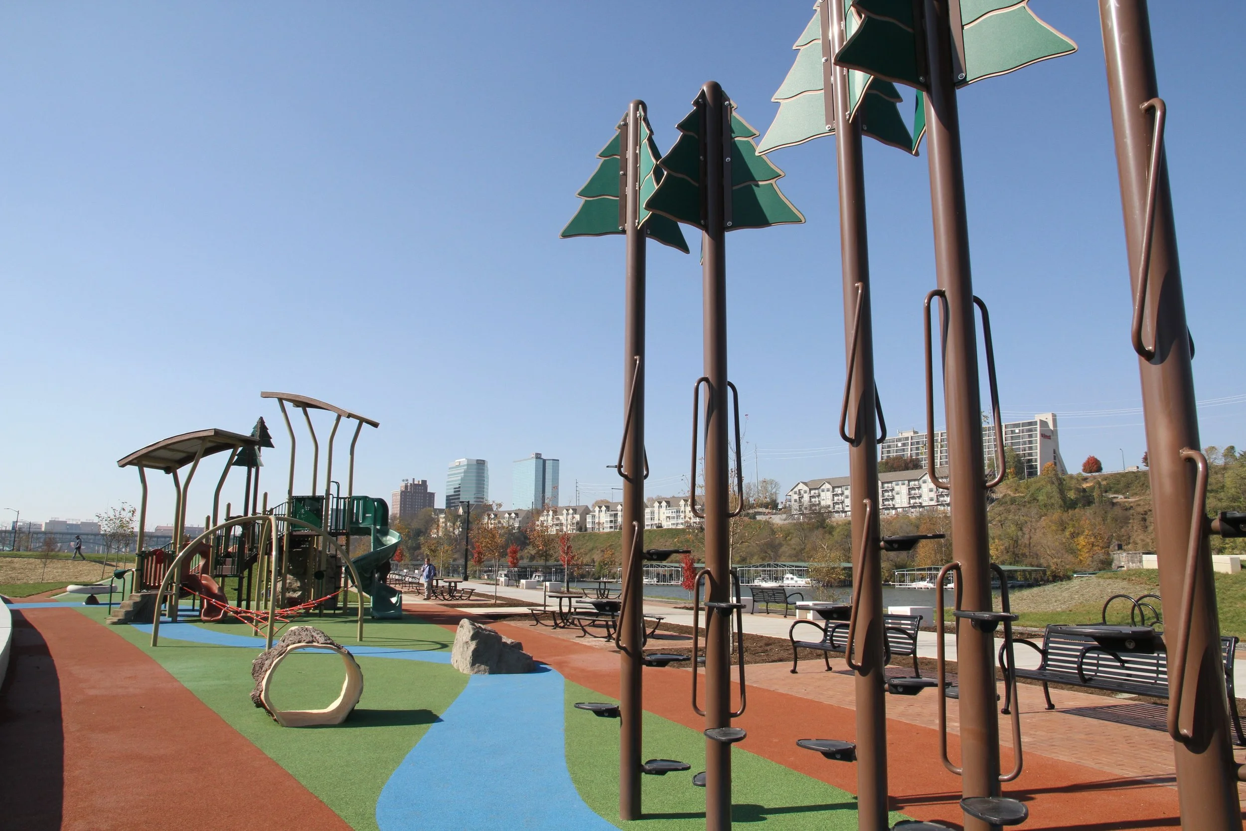 Children's playground with climbing structures, slides, and a colorful rubberized surface near a waterway with city buildings in the background.