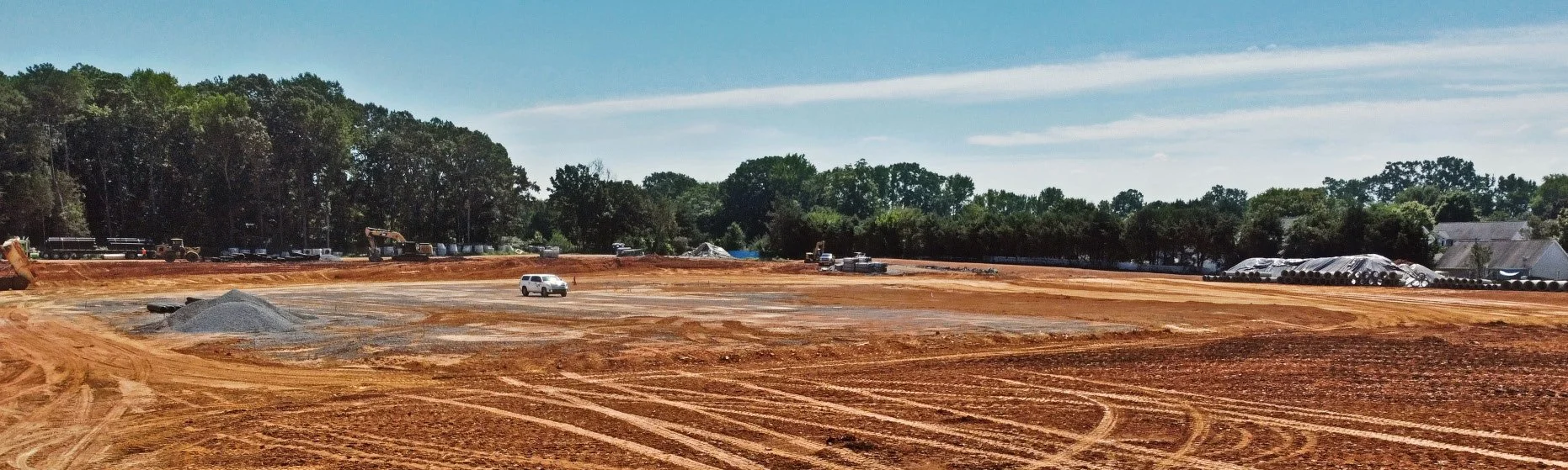 Construction site with cleared land, construction vehicles, and rows of pipes, trees in the background, and a blue sky with clouds.