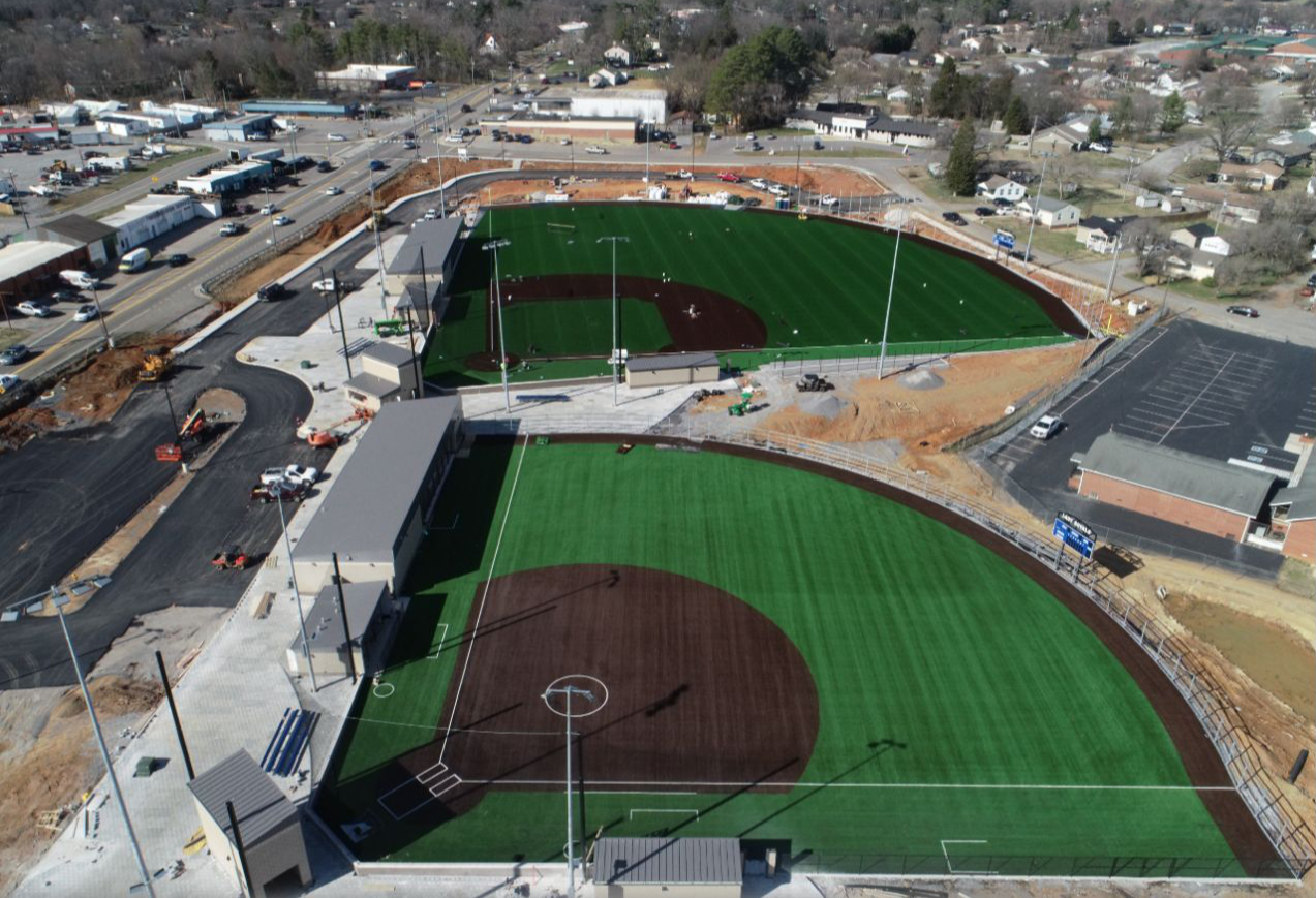 Aerial view of a baseball field under construction with green and brown turf, surrounding construction site, adjacent parking lot, and nearby buildings and streets.