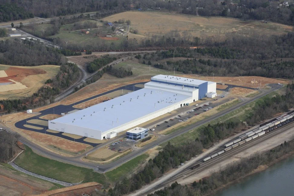 A large, white industrial or warehouse building surrounded by parking lots, train tracks with freight trains, and hills with sparse trees in the background.