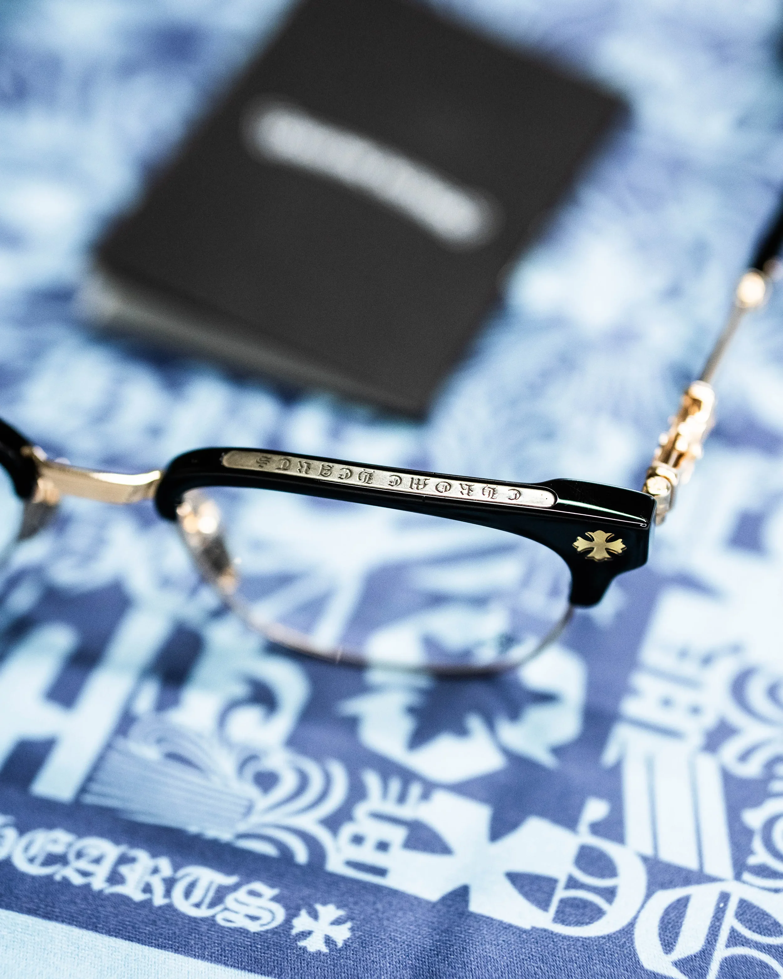 Close-up of designer sunglasses with a metal logo on the arm, resting on a blue fabric with printed text and a snowflake graphic.