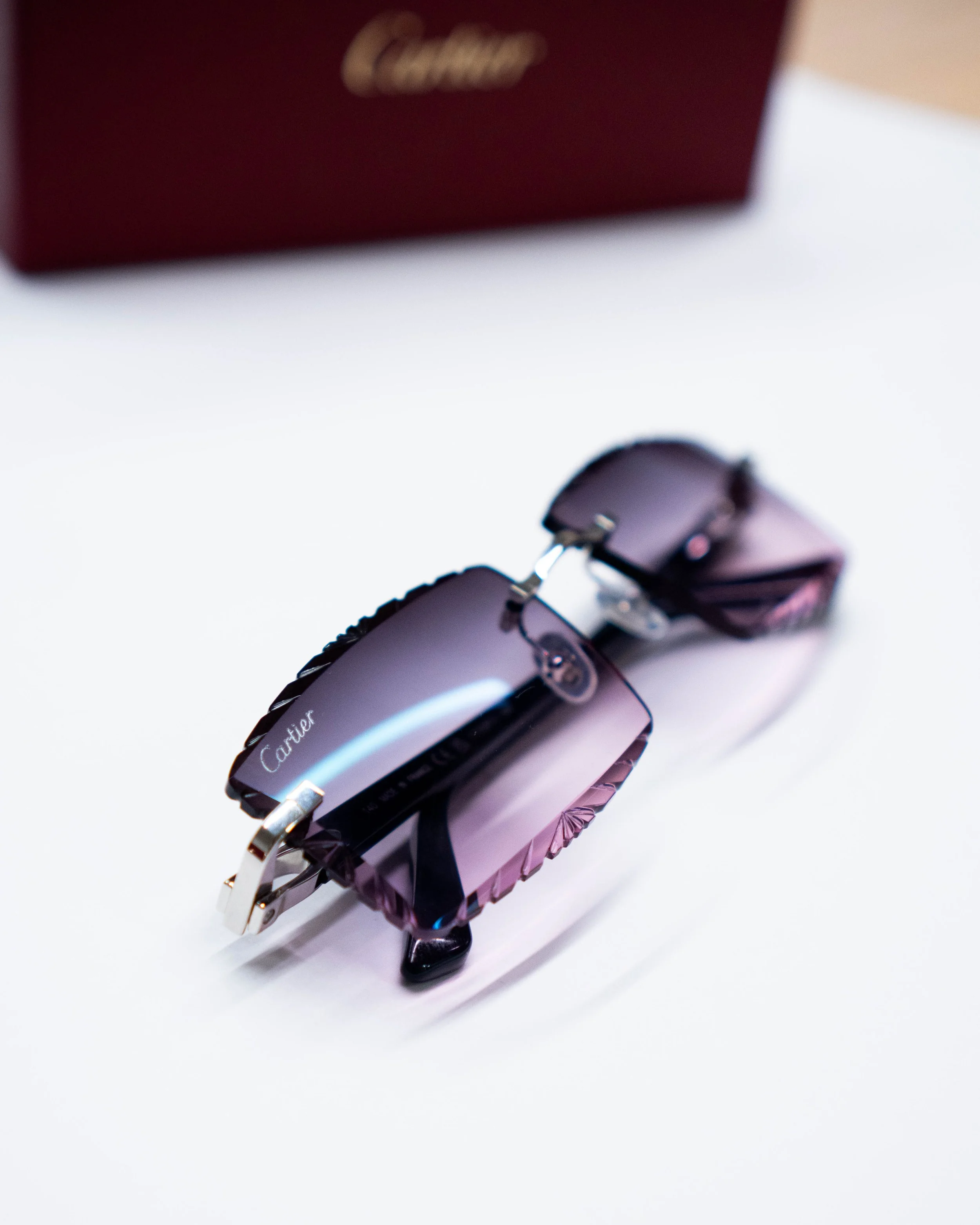 A close-up of a purple wristwatch with a jewel-encrusted band, resting on a white surface, with a dark red Cartier box blurred in the background.
