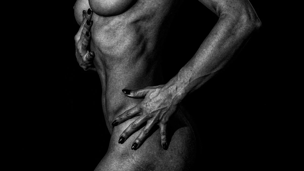 Black and white photo of a naked woman with her hand on her belly and another hand on their thigh, emphasizing muscle tone and body contours, against a dark background. Nude Art Photography by Vlad Goth.