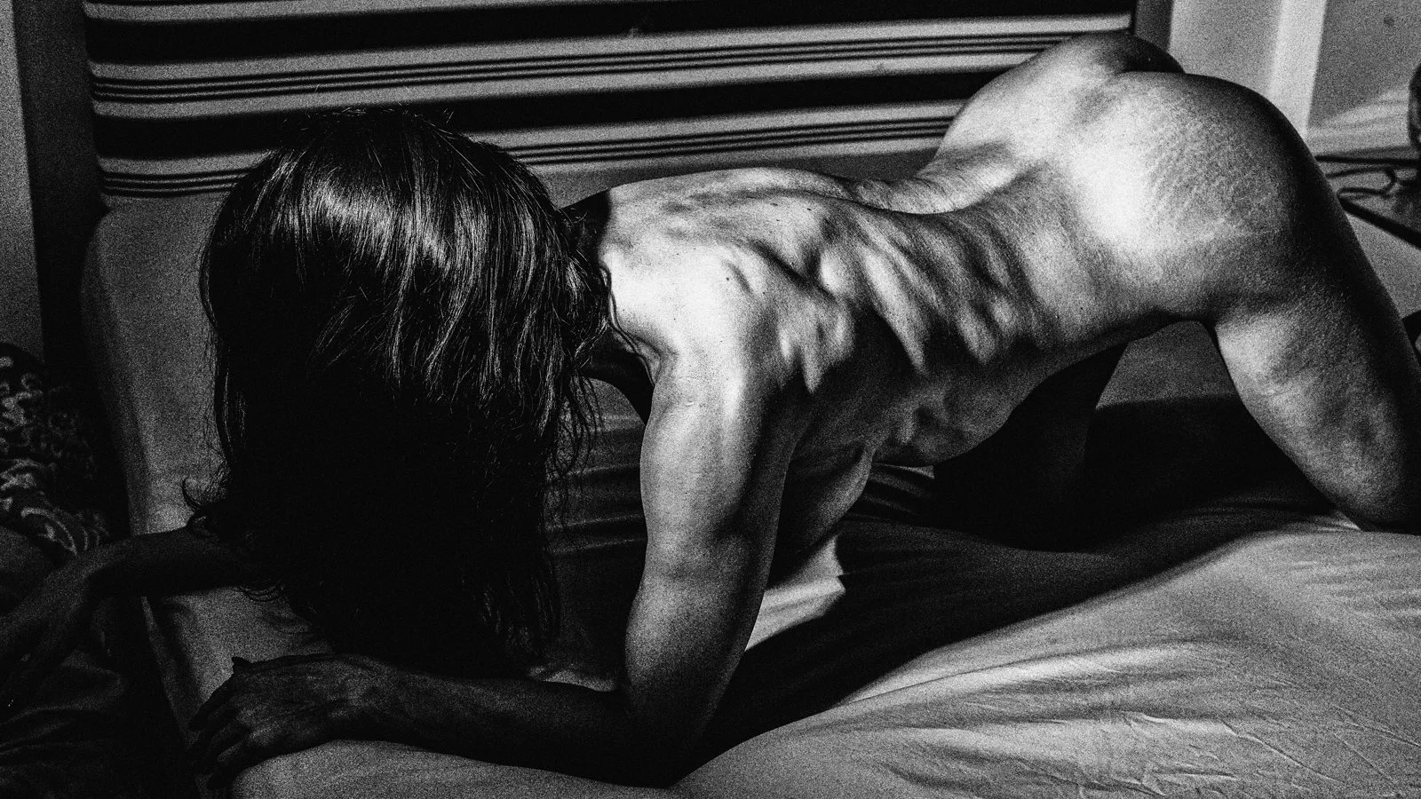 Black and white photo of a woman with long dark hair, on her legs and hands on a bed, with her head turned downward and her back exposed, with back muscle emphasised. Nude Art Photography by Vlad Goth.