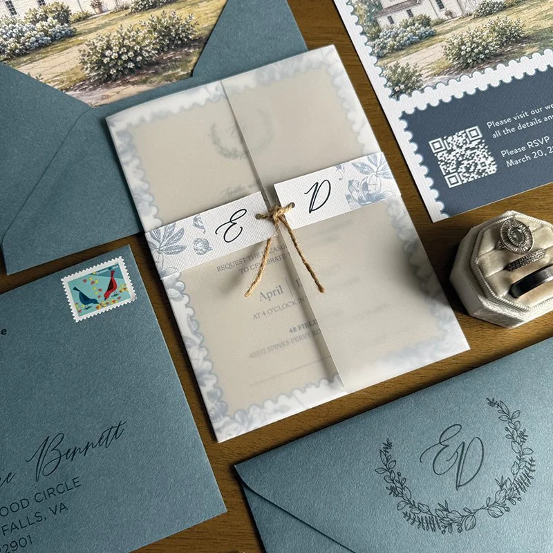 Wedding invitation suite with blue envelopes, RSVP card, and floral details, along with a ring in a velvet box, on a wooden table.
