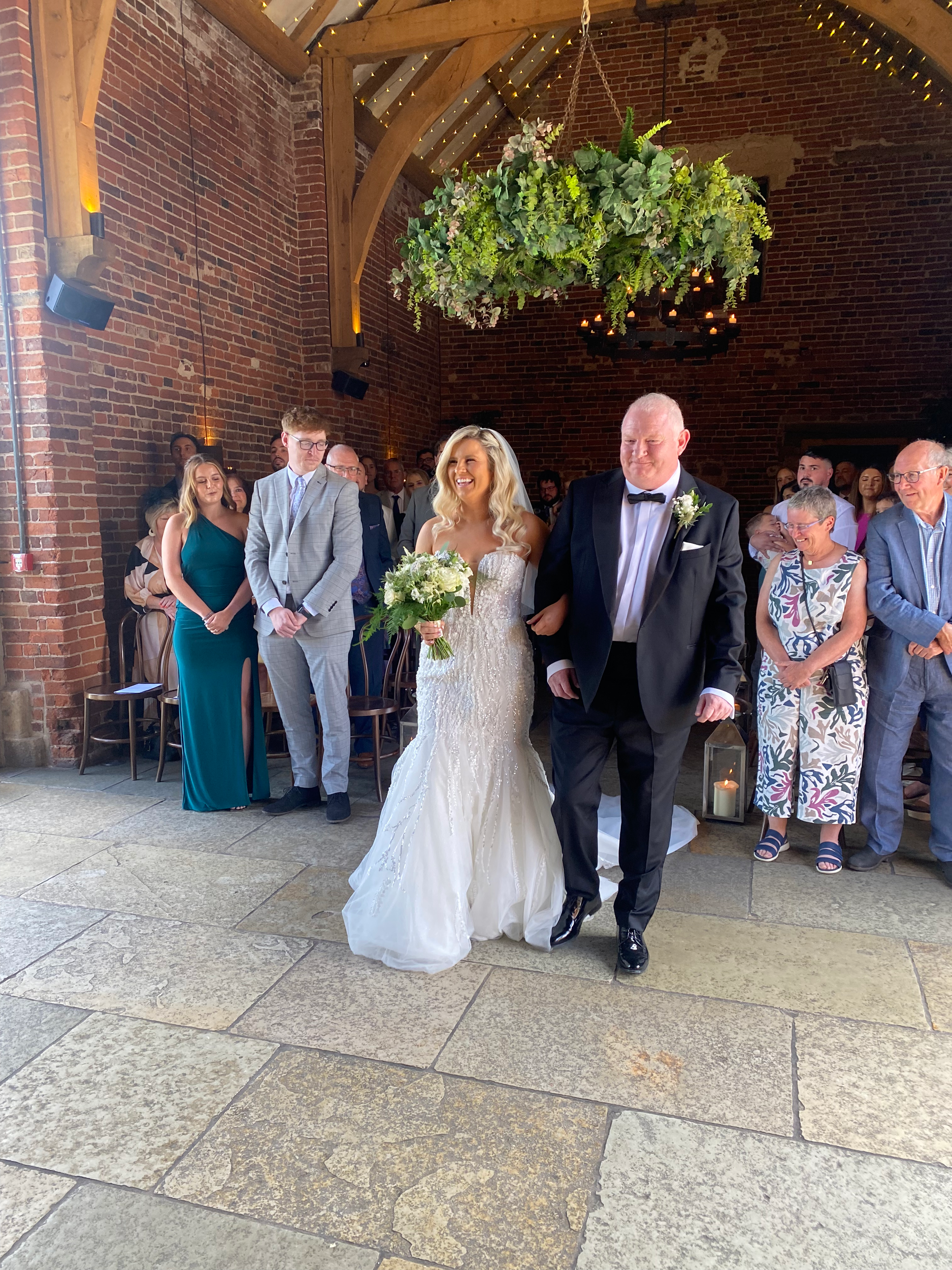 A wedding ceremony with a bride in a white dress and groom in a tuxedo, surrounded by guests, in an indoor venue with brick walls and decorative lighting, during daytime.