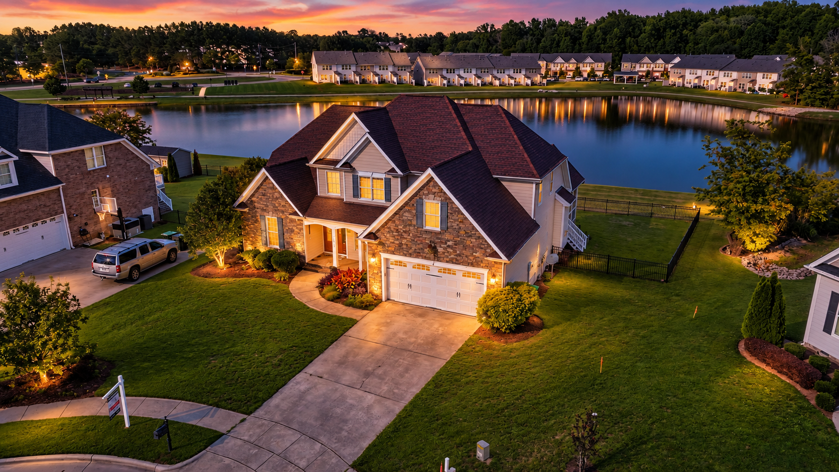 A house with stone and siding exterior surrounded by well-maintained grass and trees, illuminated by exterior lights at dusk. A driveway leads to a garage, and a lake with reflections of nearby houses and trees is in the background.