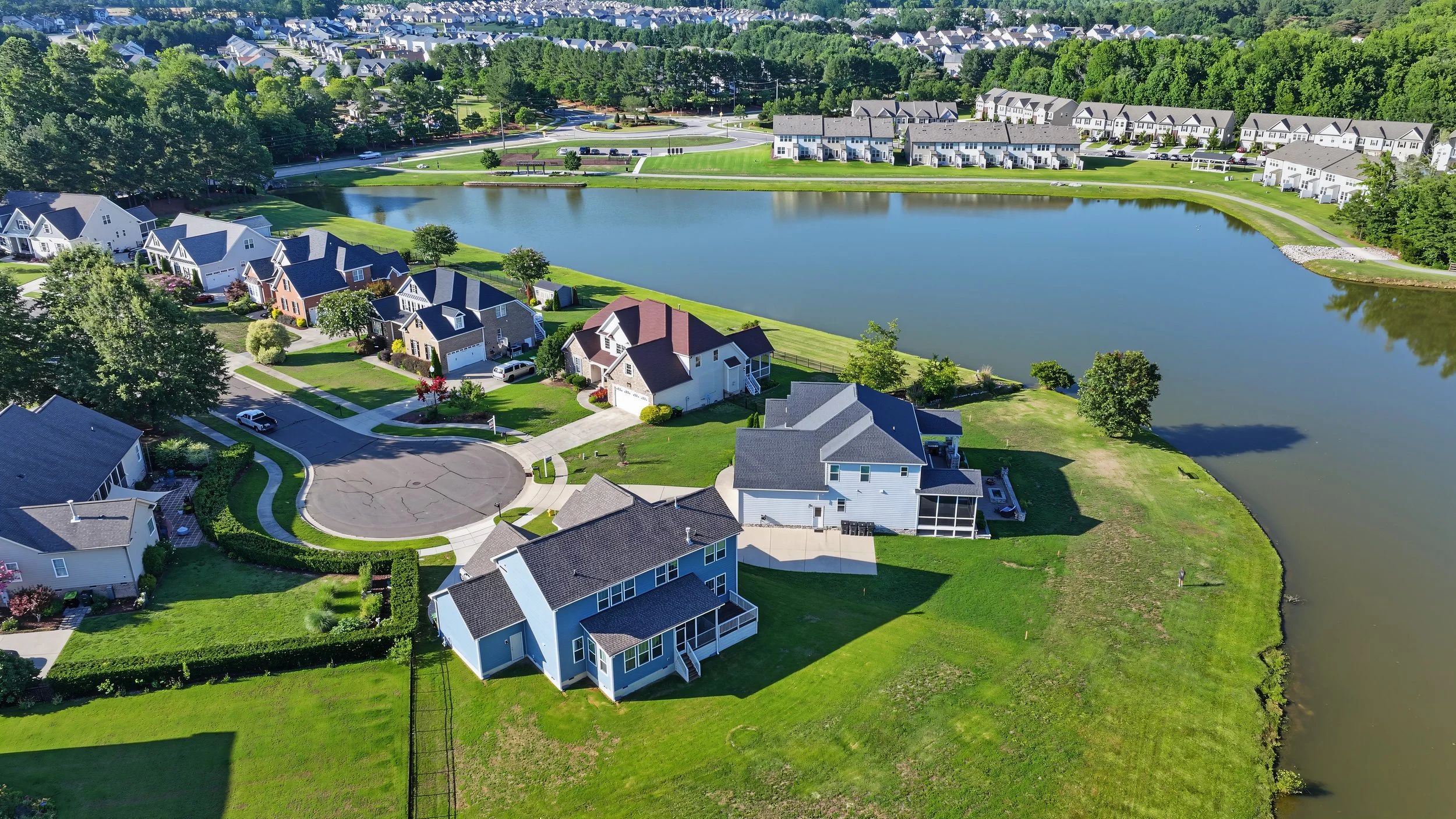 Aerial view of a suburban neighborhood with houses, a pond, and green lawns on a sunny day.