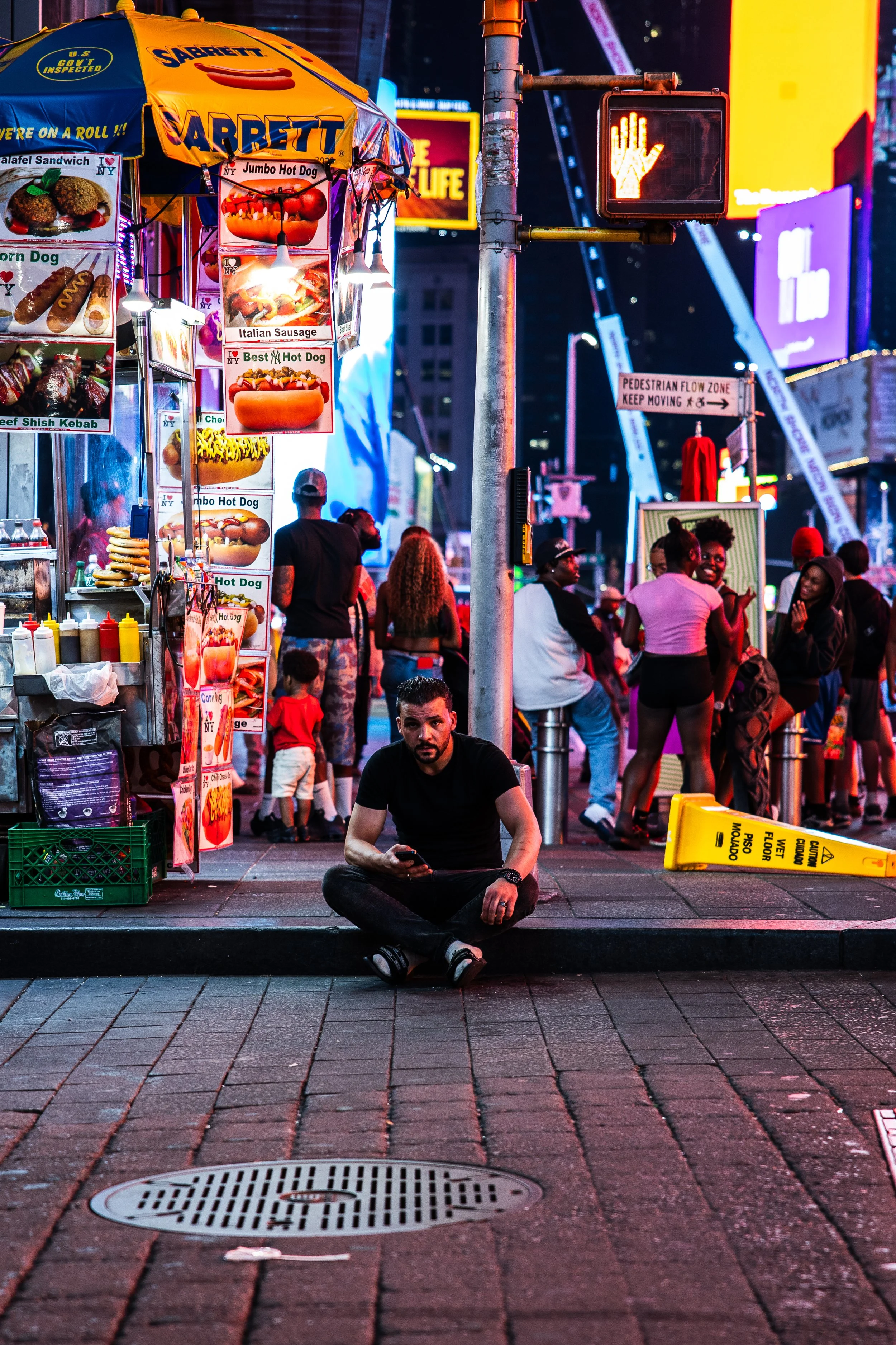 Nighttime scene in a city with food stands, neon signs, and people walking around. A man is sitting on the sidewalk using his phone, with a yellow wet floor caution sign nearby.