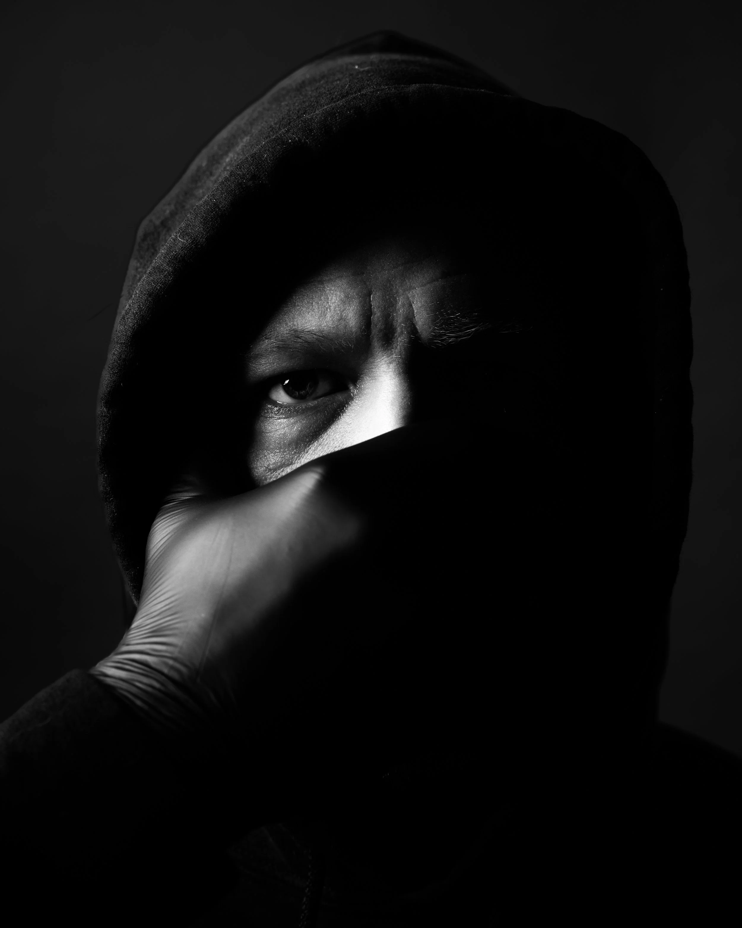 Close-up black and white photograph of a person's face partially covered by a hoodie, revealing one eye and a gloved hand holding the hoodie near the face.