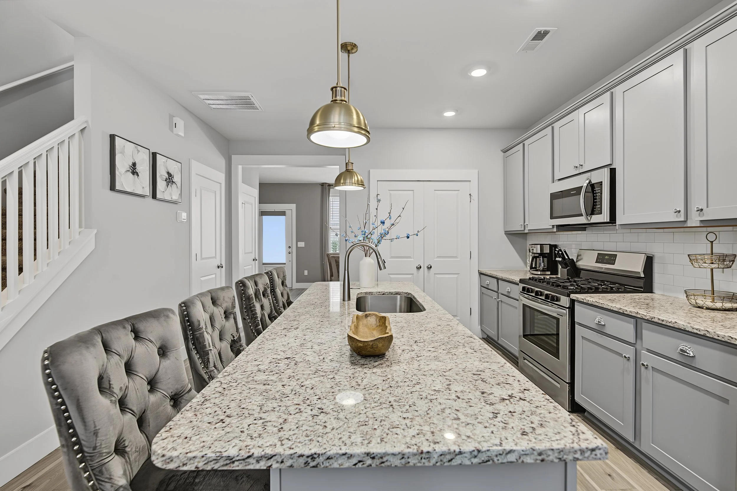 Modern kitchen with gray cabinets, stainless steel appliances, granite countertops, and a beige upholstered dining chair with silver nailhead trim.