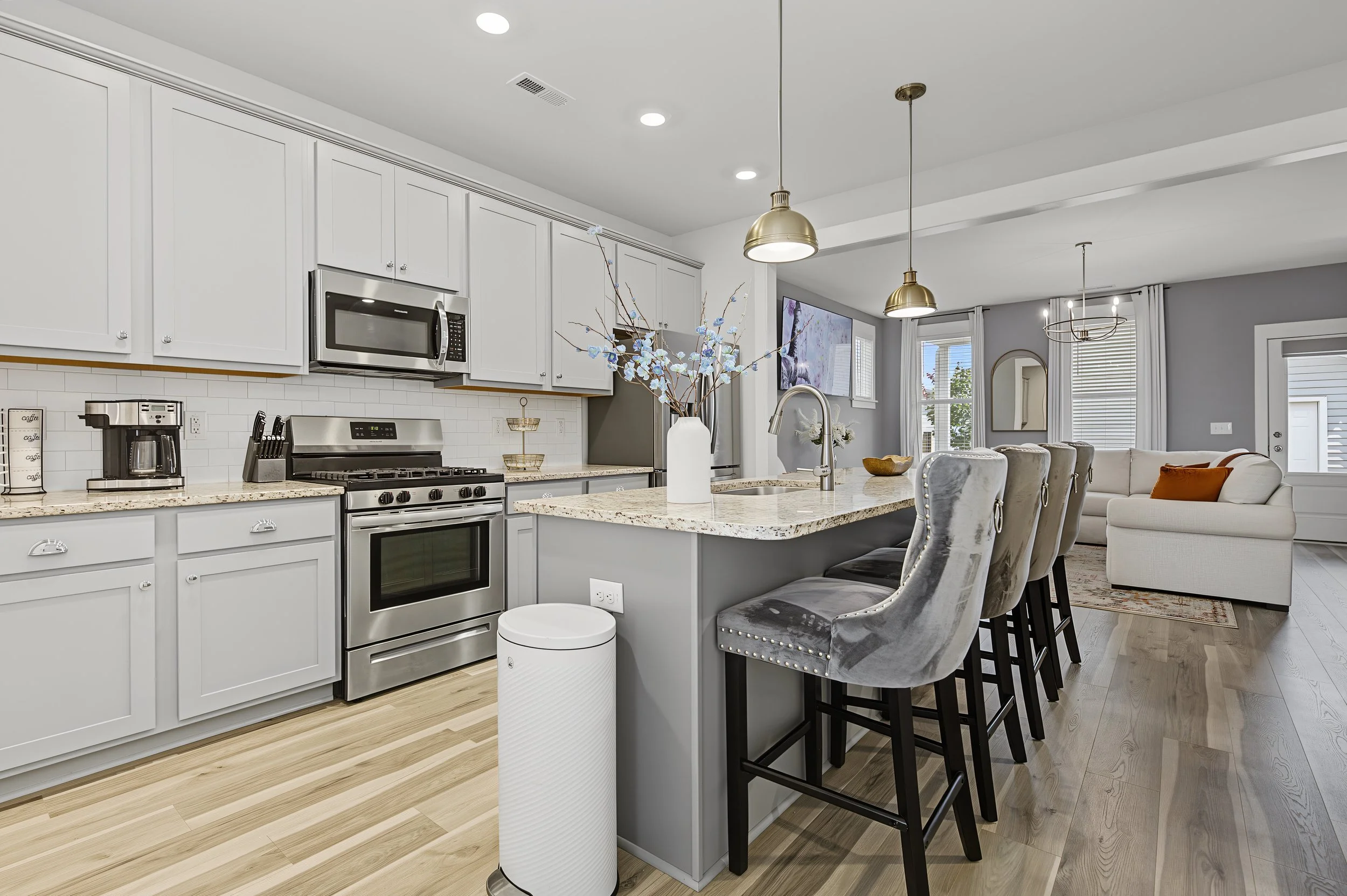 Modern kitchen with white cabinets, stainless steel appliances, a granite island with a vase of blue flowers, and gray velvet chairs at the counter. Open living space with large windows, curtains, and a television.
