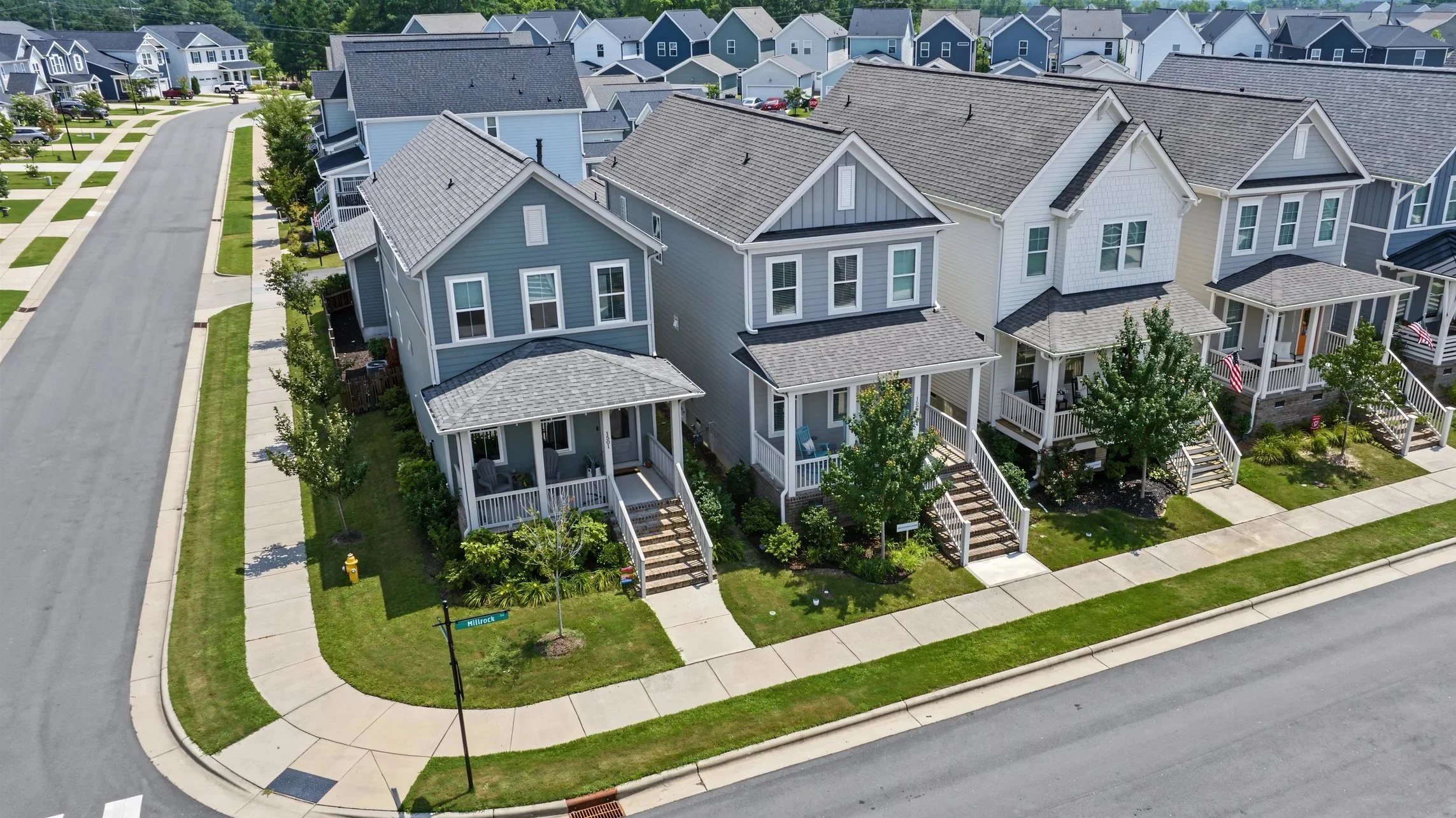 Aerial view of a modern residential neighborhood with multi-story houses, manicured lawns, and sidewalks, situated along a curved street.