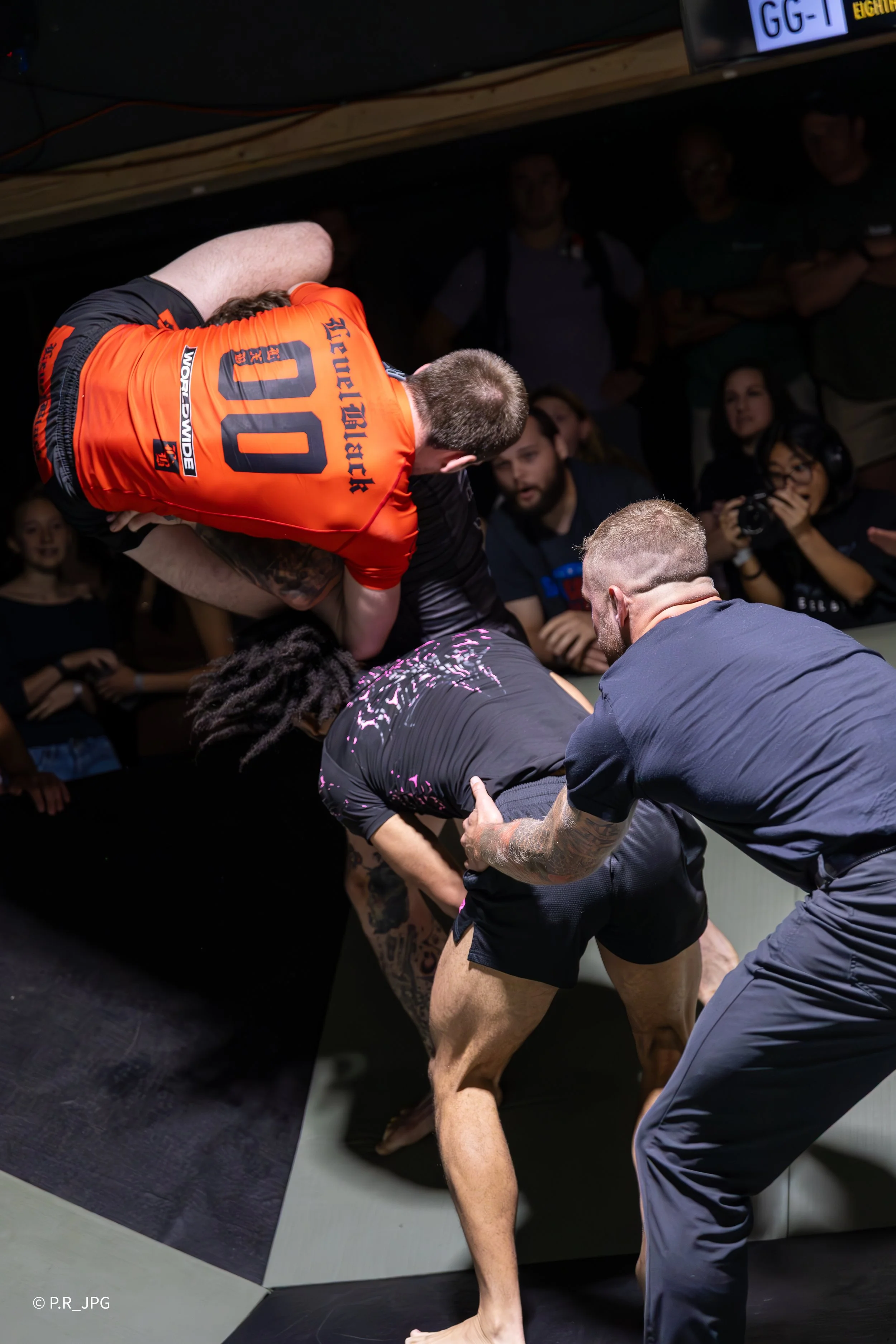 Two men practicing Brazilian Jiu-Jitsu or grappling on a mat in a training gym, with spectators watching in the background.