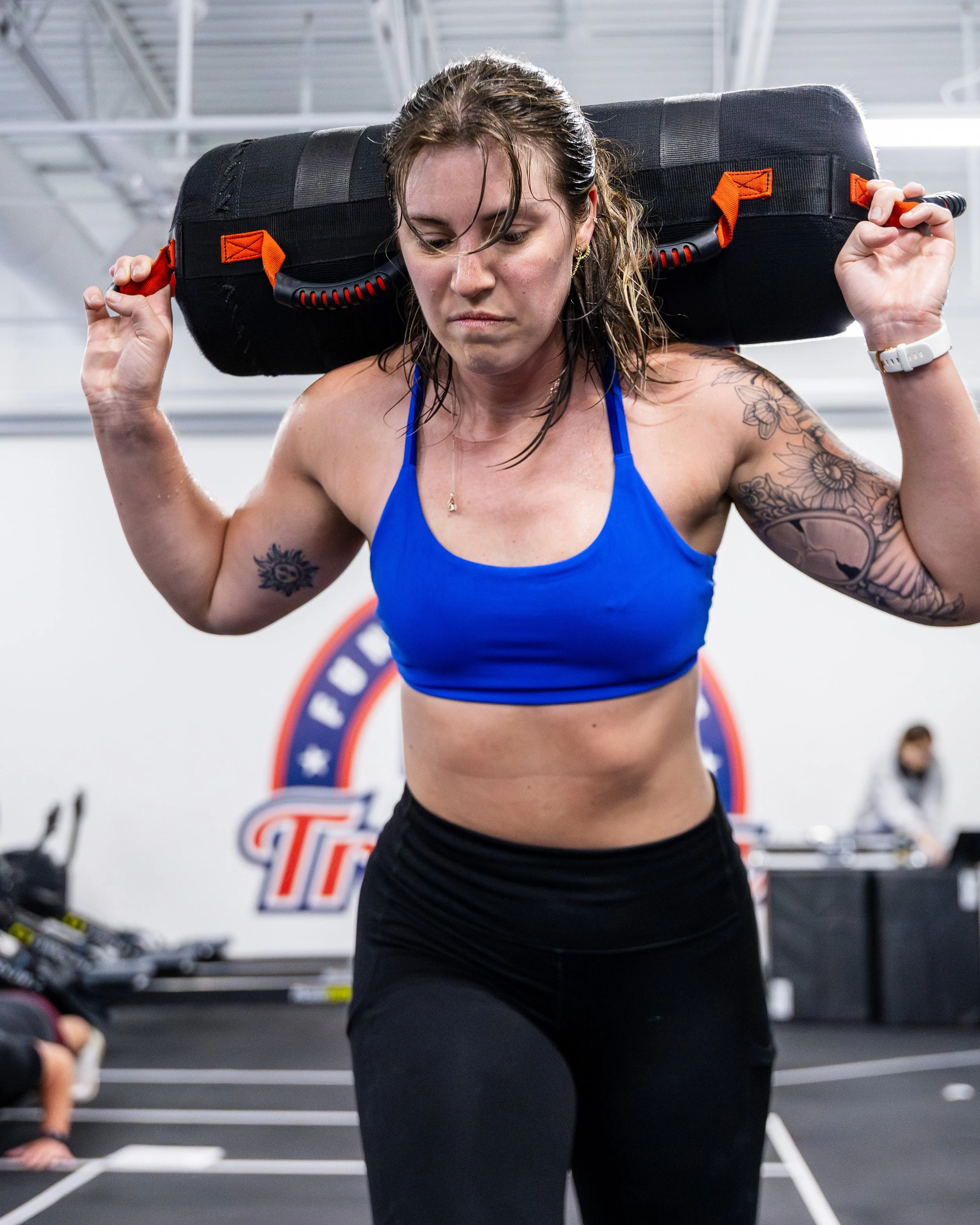 A woman in a blue sports top and black leggings acts as if she's carrying a heavy object on her shoulders in a gym.