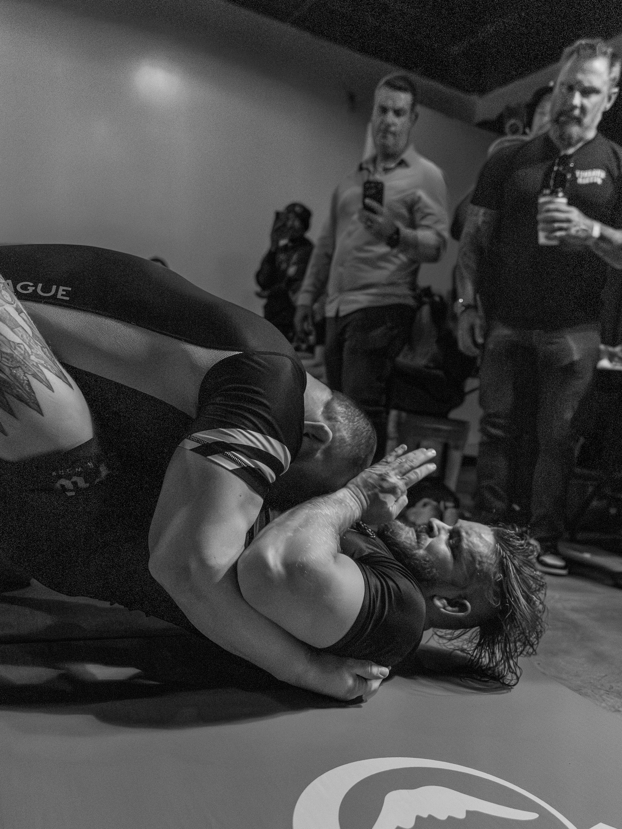 A woman in a black shirt and leggings is lying on the floor, holding her head with both hands in pain or distress. Several people, including a man in a polo shirt and a man with glasses in a button-up shirt, are standing around her, looking concerned or interested. The setting appears to be an indoor gym or training area.