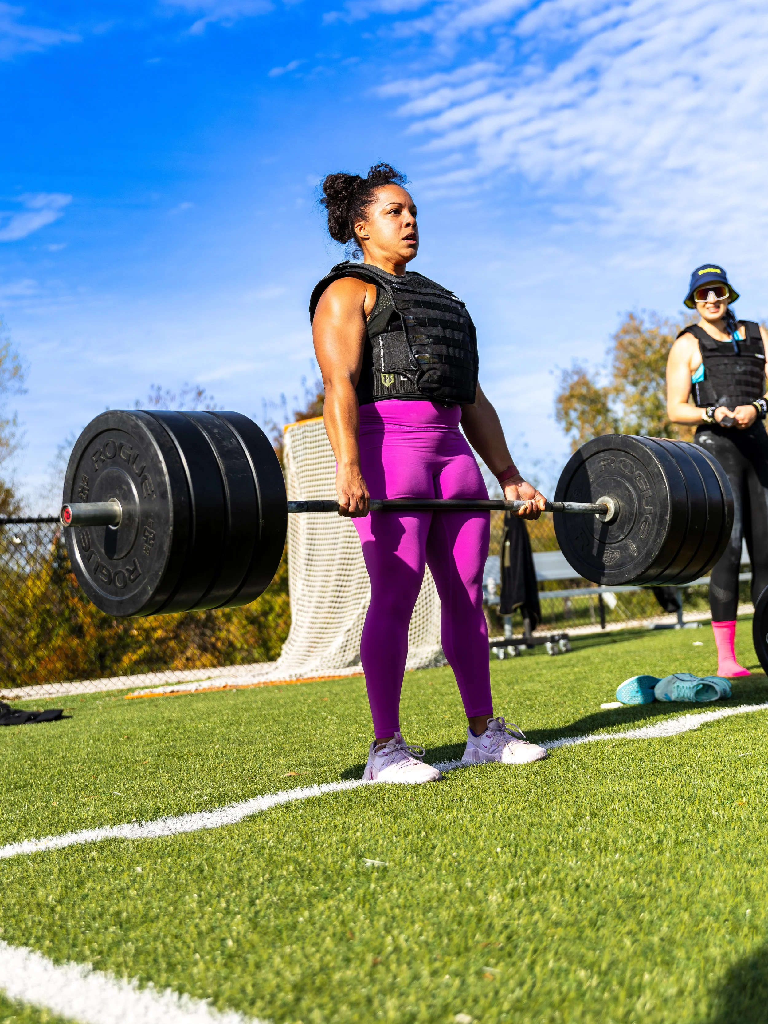 Women in athletic wear lifting a barbell with large weights outdoors on a sunny day.