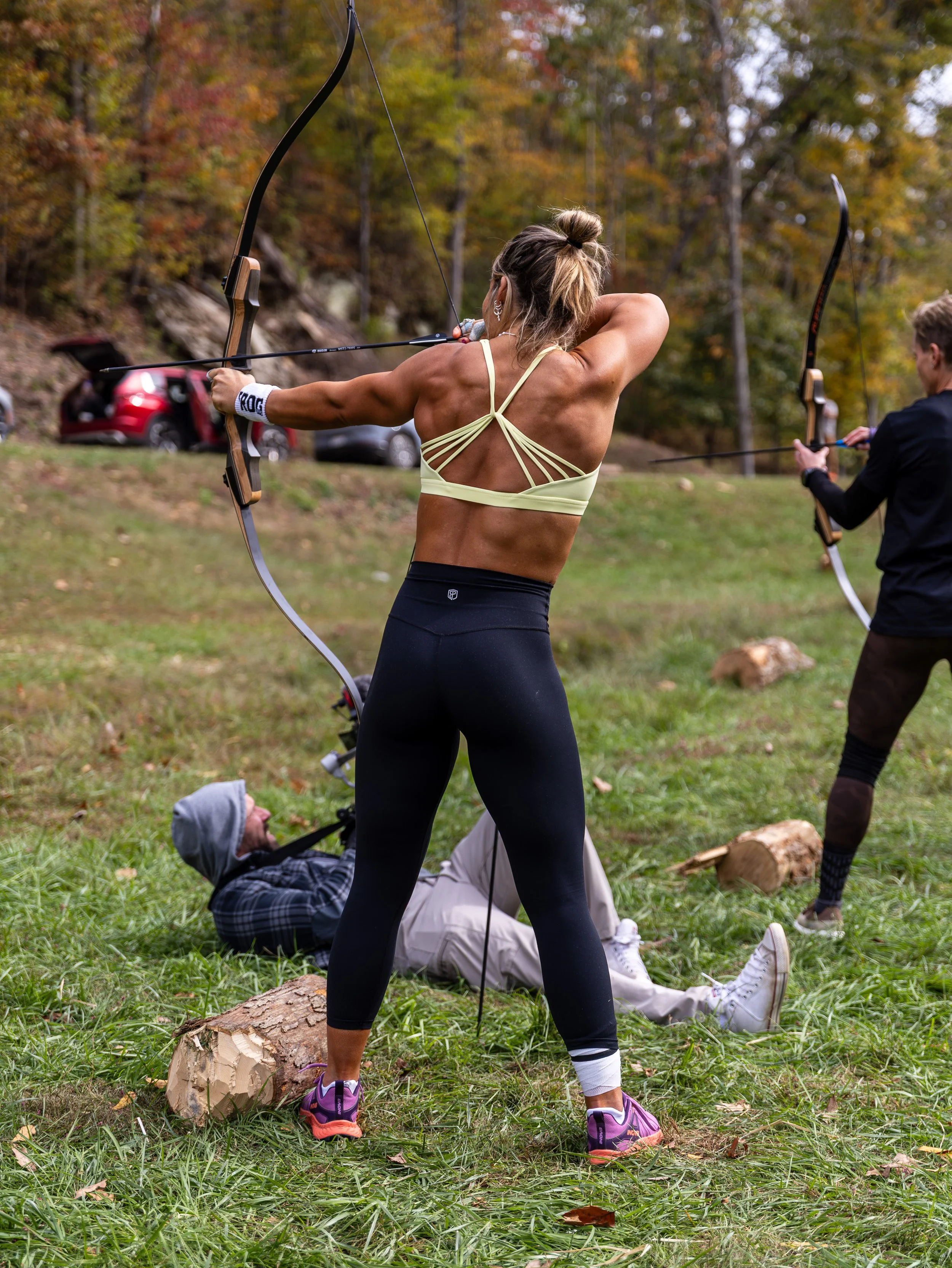 Woman practicing archery outdoors in a park with autumn trees, a fallen log on the grass, and another person in the background also aiming an arrow.