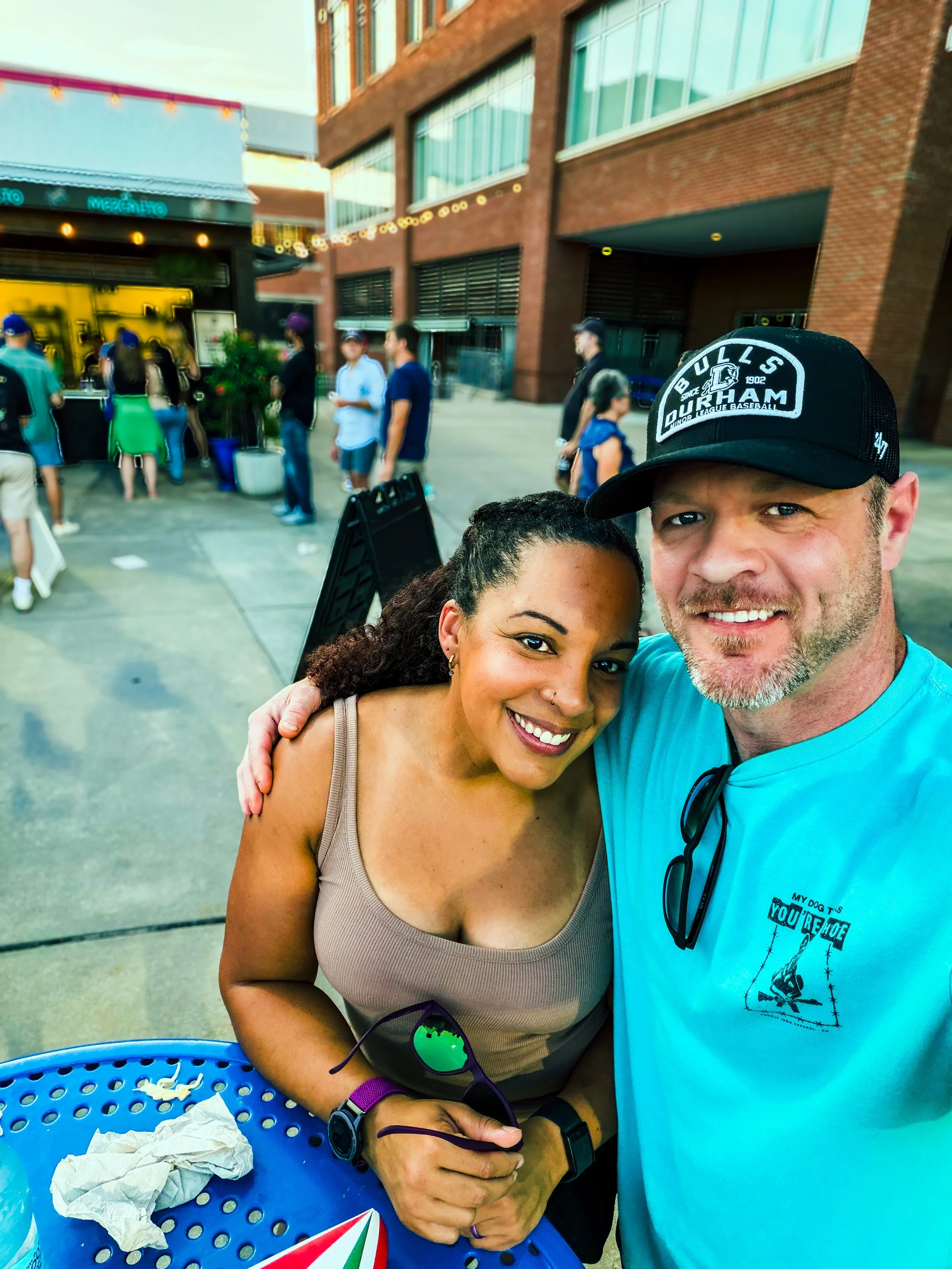 Close-up of a woman and man taking a selfie outdoors with a brick building and people in line in the background.