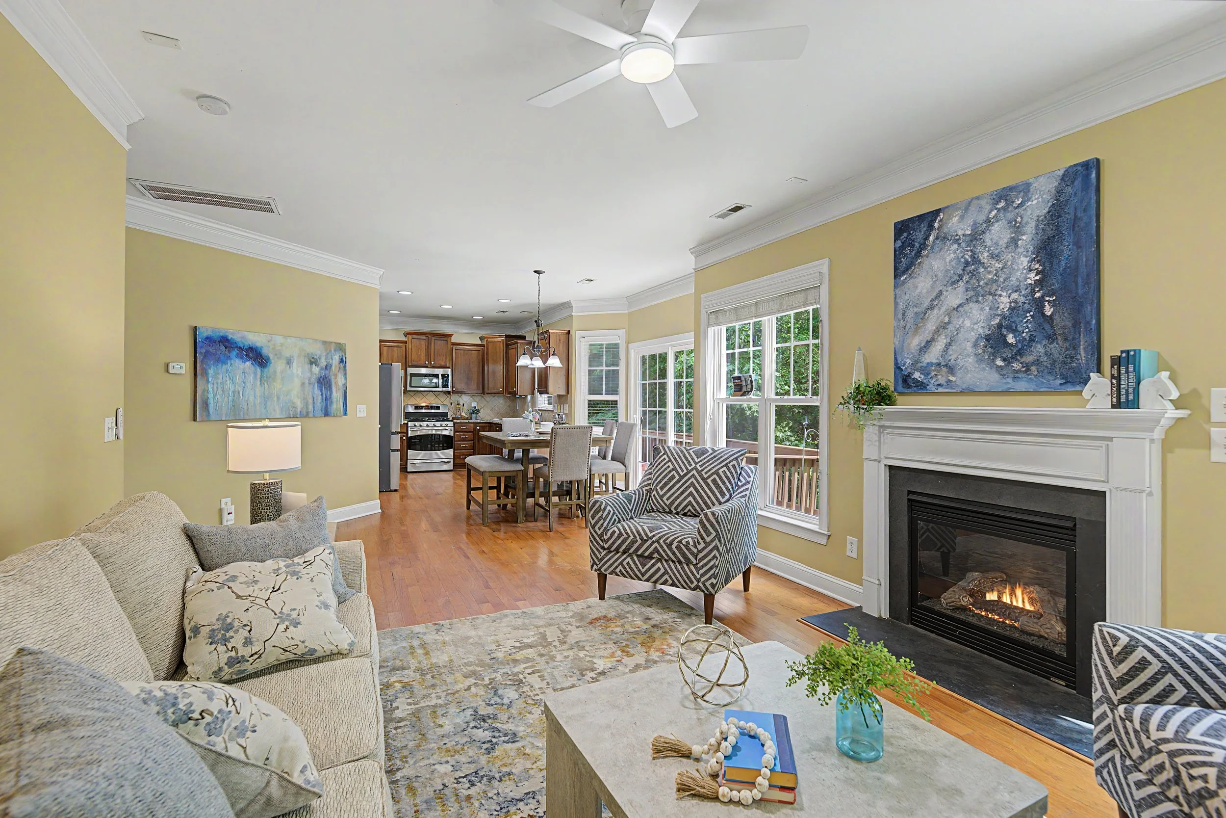 Living room with beige sofa, patterned chairs, and a marble coffee table with decorative objects. Open kitchen with wooden cabinets, stainless steel appliances, and dining table in background. Bright windows with green view, white ceiling fan, and a fireplace with white mantel. Artworks and books decorates the space.
