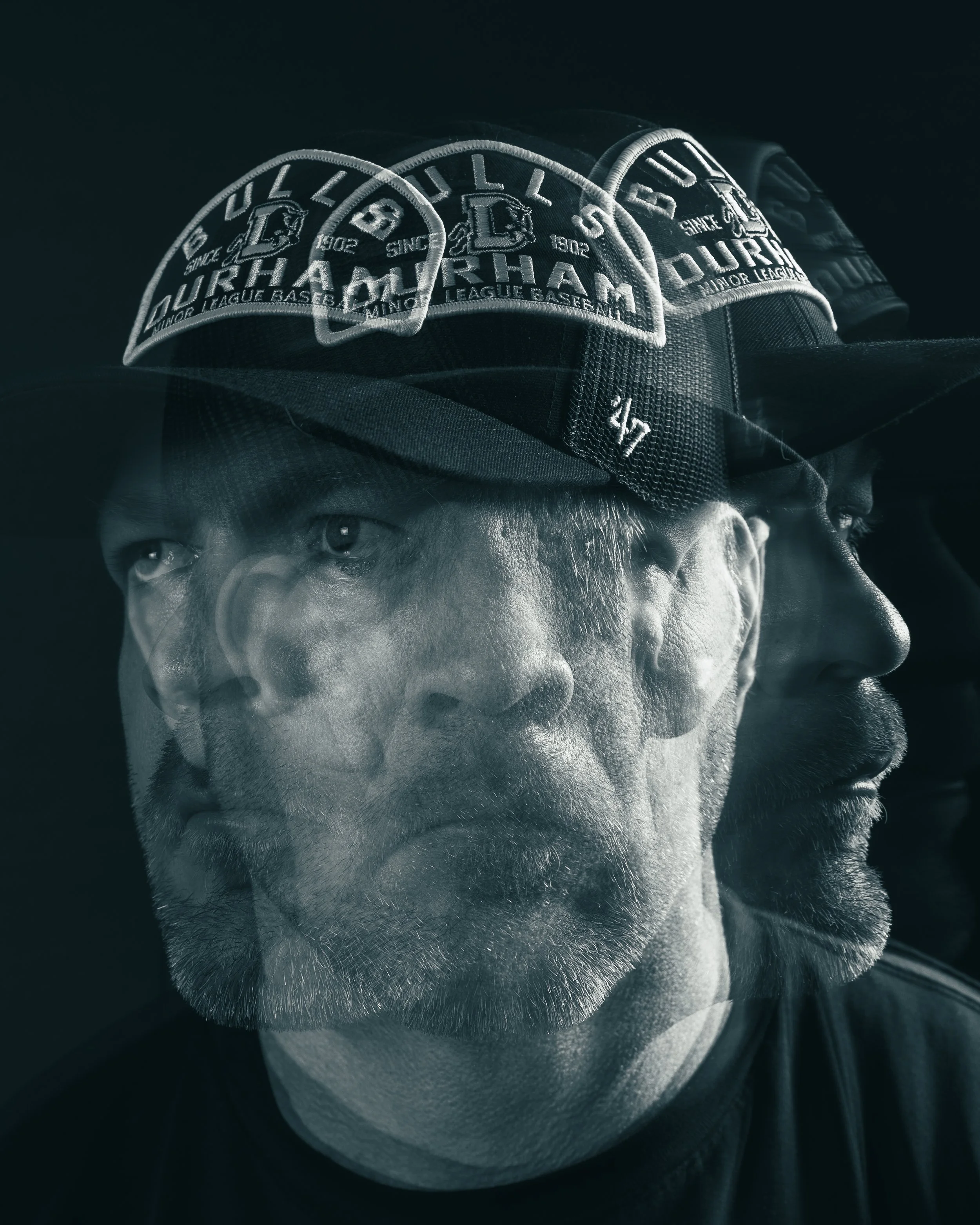 Close-up of a person's face and baseball cap featuring the Durham Bulls logo, a minor league baseball team, with the person looking to the side and the image captured in monochrome.