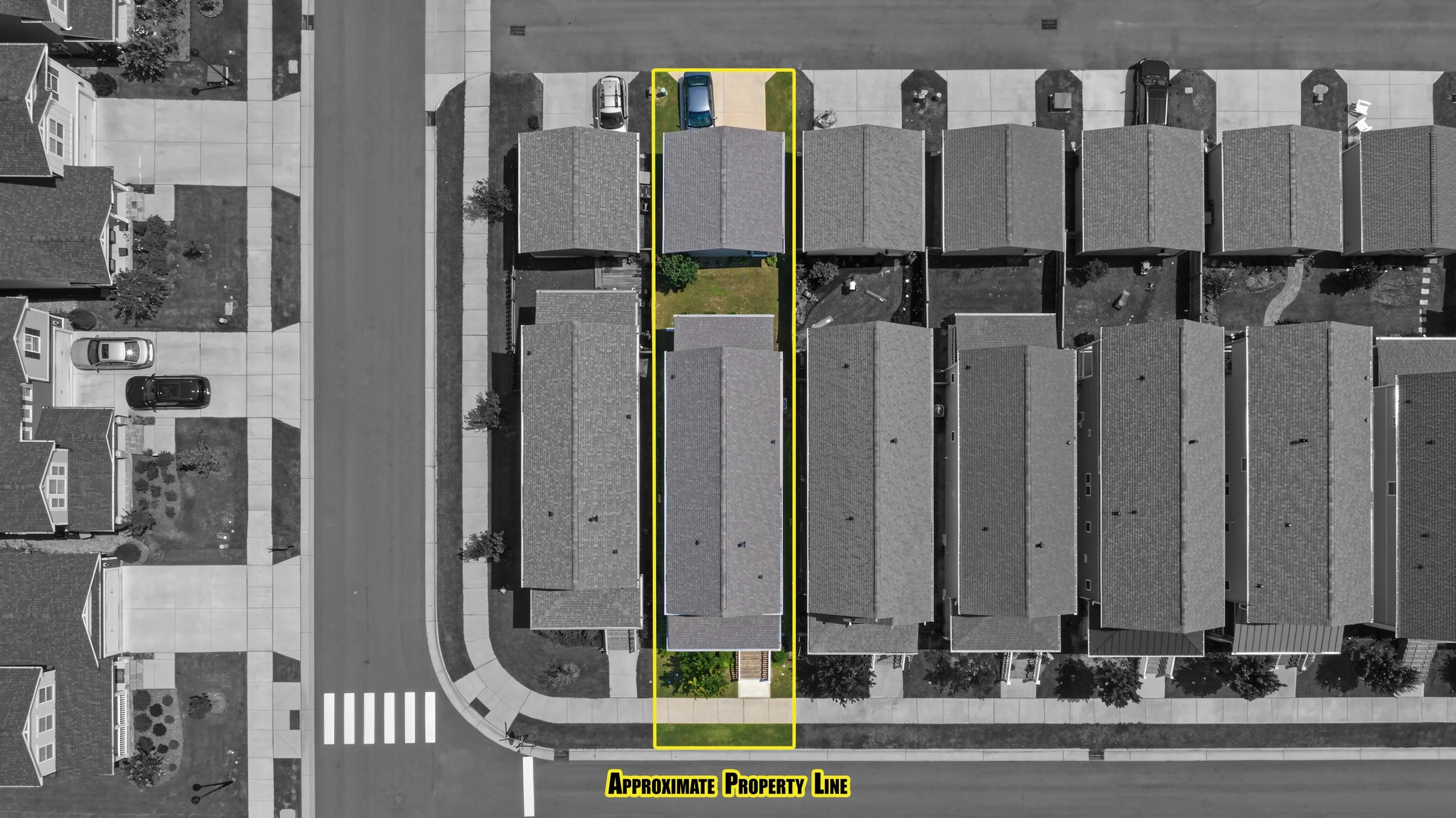 An aerial view of a residential neighborhood showing multiple houses, parked cars, and a sidewalk. A property outlined in yellow has a backyard with grass, small trees, and a driveway. The approximate property line is marked with a yellow line and labeled at the bottom.