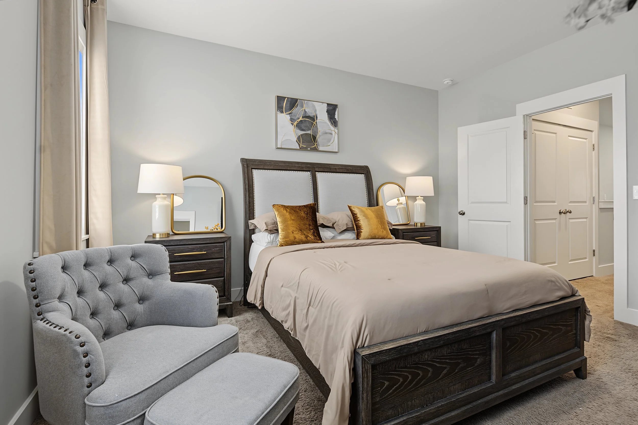 A bedroom with a large bed, two matching nightstands with lamps, a decorative painting above the bed, a gray upholstered armchair, and a set of white double doors. Real estate photography.