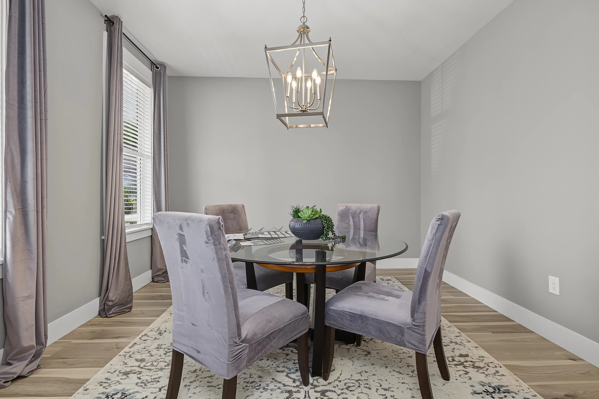 A dining room with a round glass table surrounded by six upholstered chairs, a chandelier hanging above, grey curtains covering the windows, and a decorative plant in a black pot on the table. The room has light-colored walls and wood flooring.