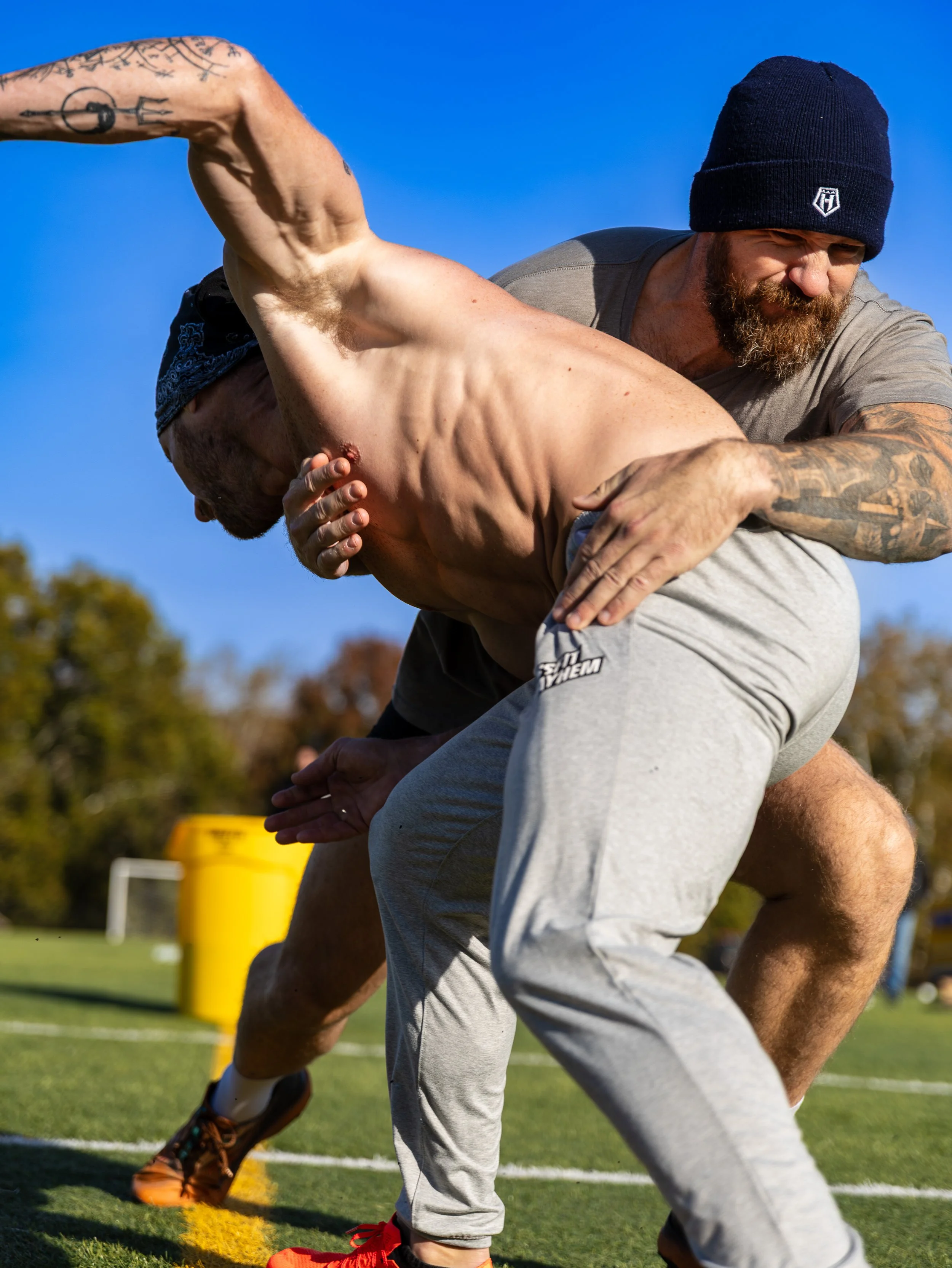 Two men engaged in a wrestling or grappling match outdoors on a grassy field, with a soccer goal and yellow trash bin in the background, under a clear blue sky. One man is holding the other from behind, and both are wearing casual athletic clothing.