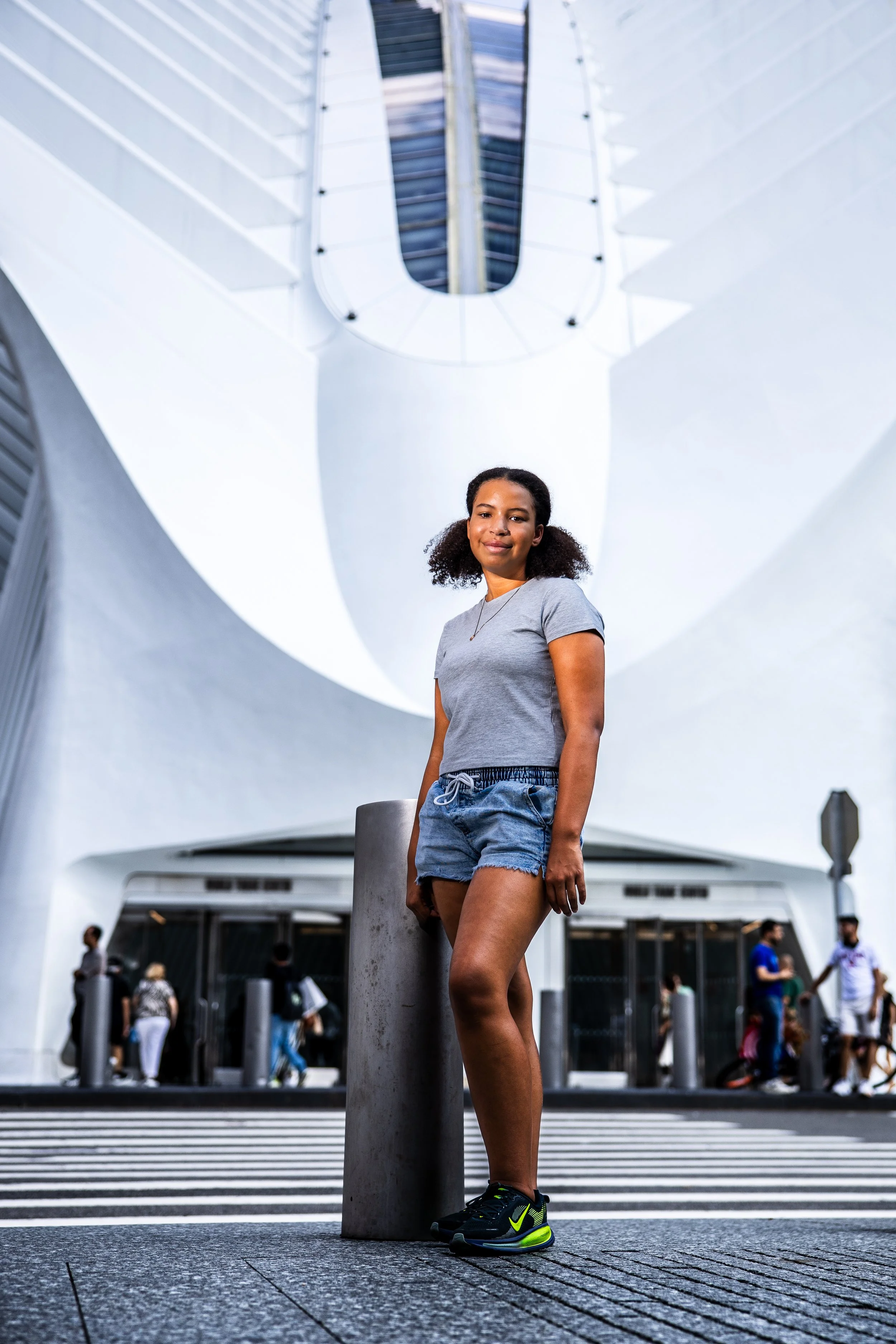 Young woman in gray t-shirt and denim shorts standing in front of modern building with people walking in background, close-up shot from below.