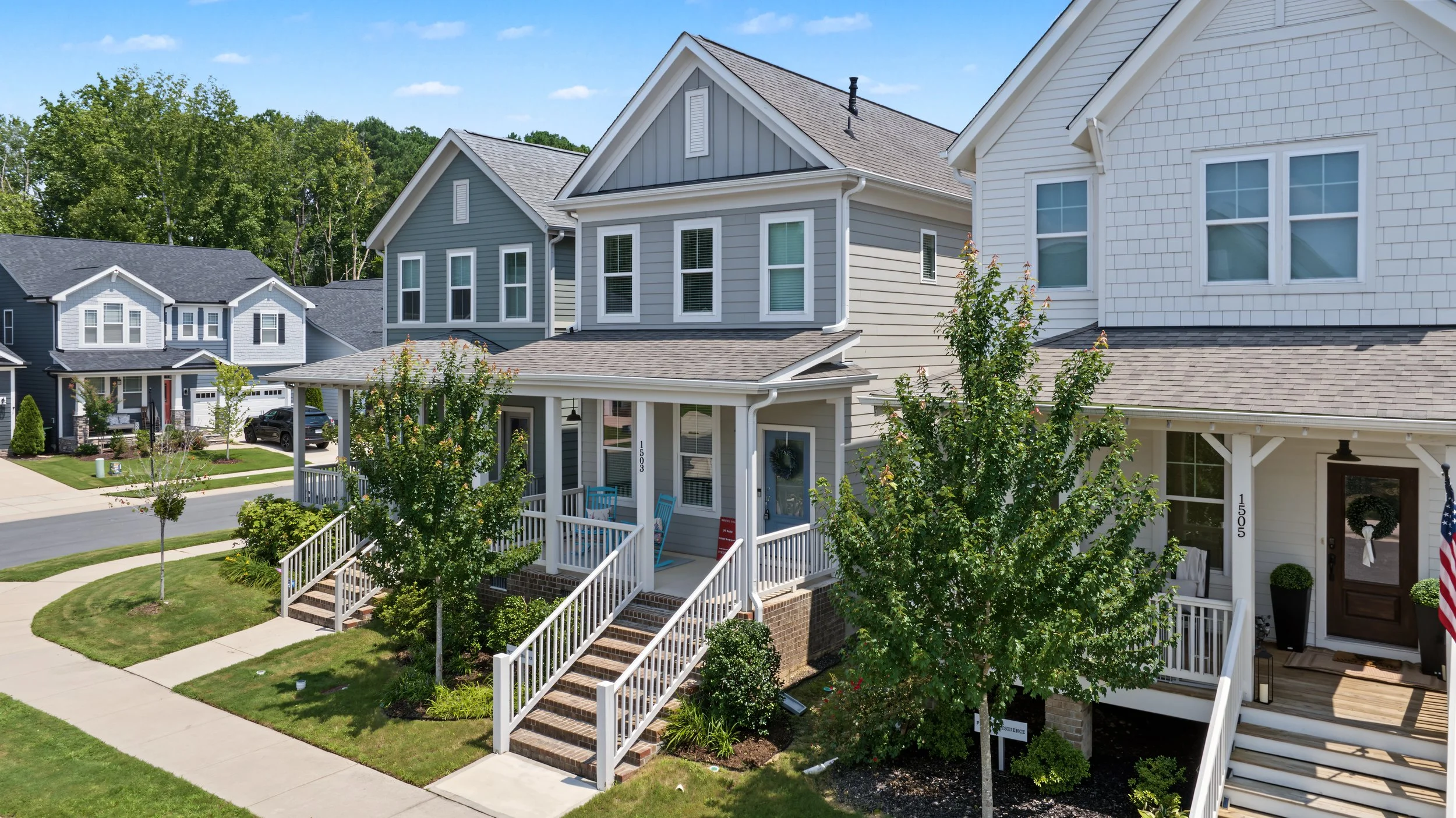 A row of modern suburban houses with front porches, white and gray siding, and well-maintained lawns under a clear blue sky.