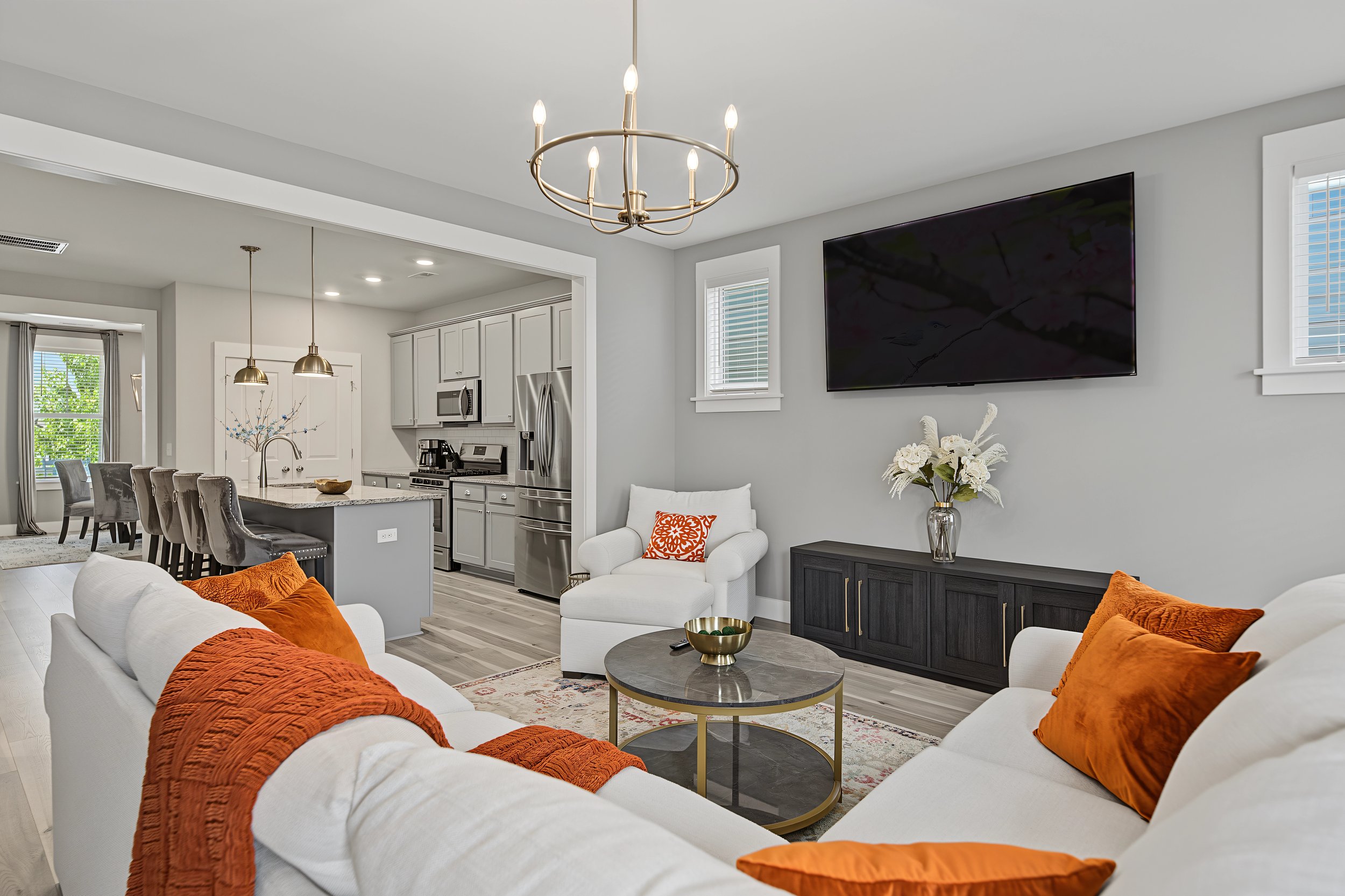Living room with white sofas, orange throws, a round coffee table, a TV on the wall, and an open kitchen with white cabinets and stainless steel appliances.