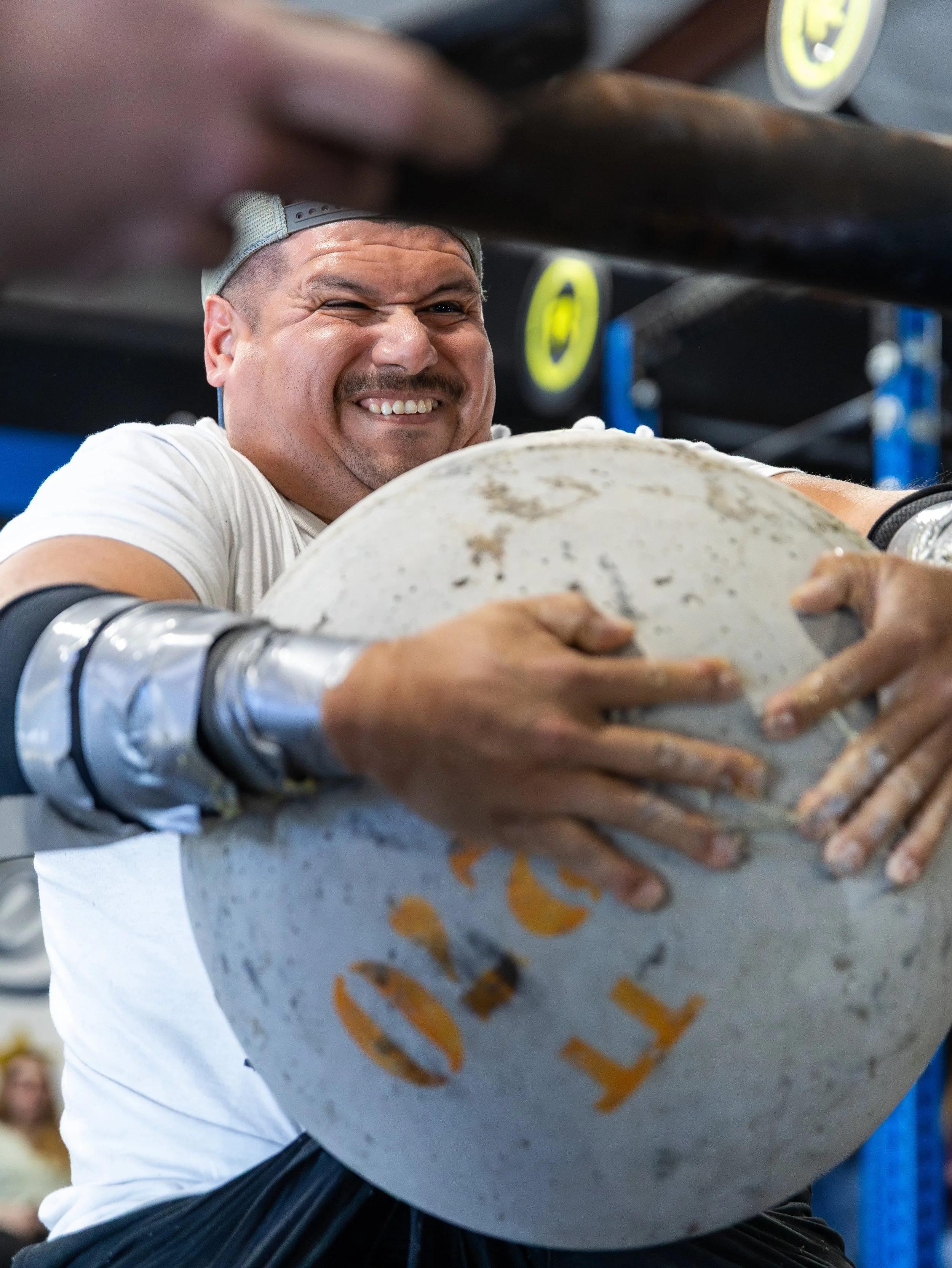 A man in a white shirt and baseball cap, smiling and winking, appears to be lifting or holding a large, worn white object with orange markings, inside a gym or fitness setting.