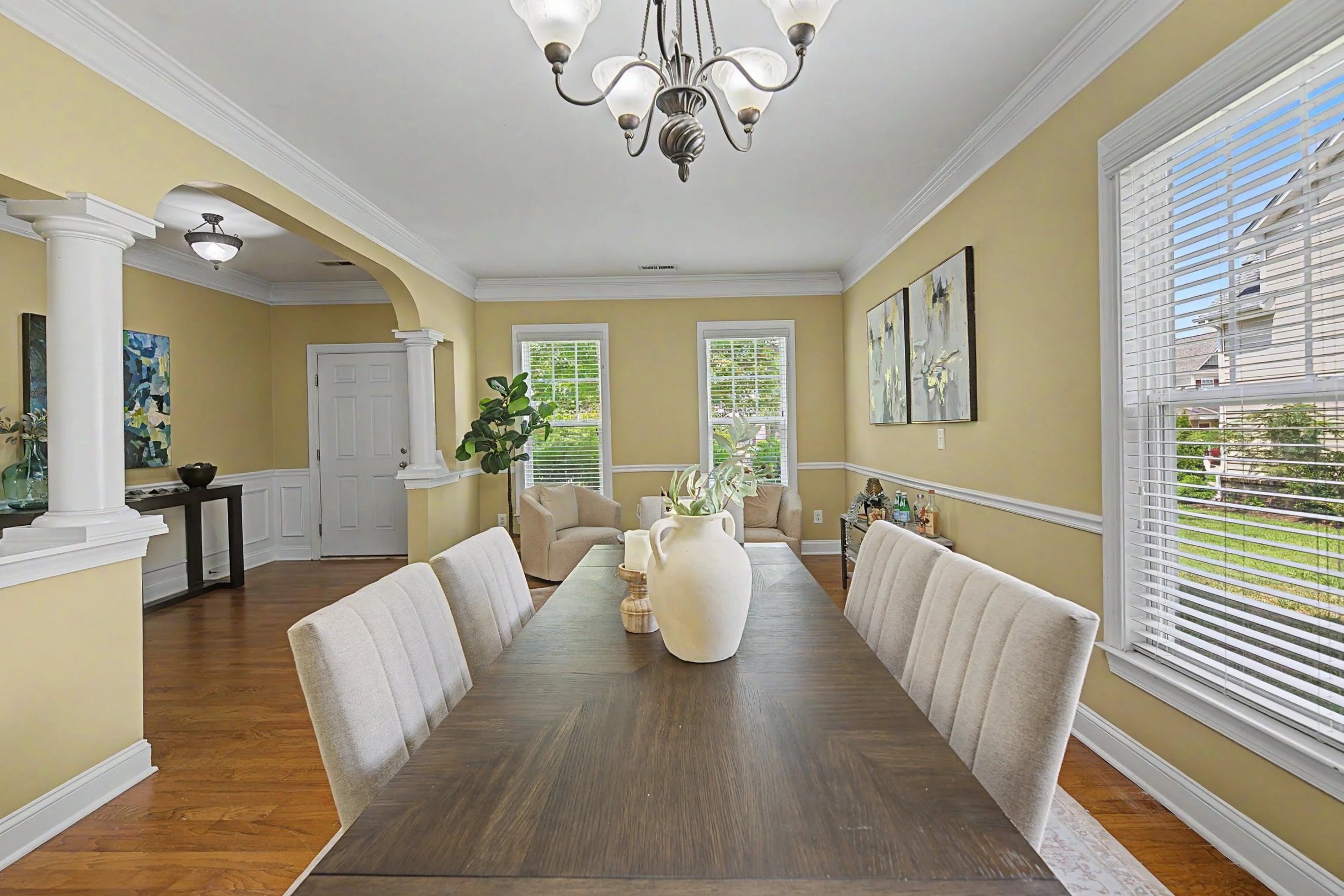 A dining room with a dark wooden table, beige upholstered chairs, decorative vases, and large windows with white blinds, yellow walls, and white crown molding.