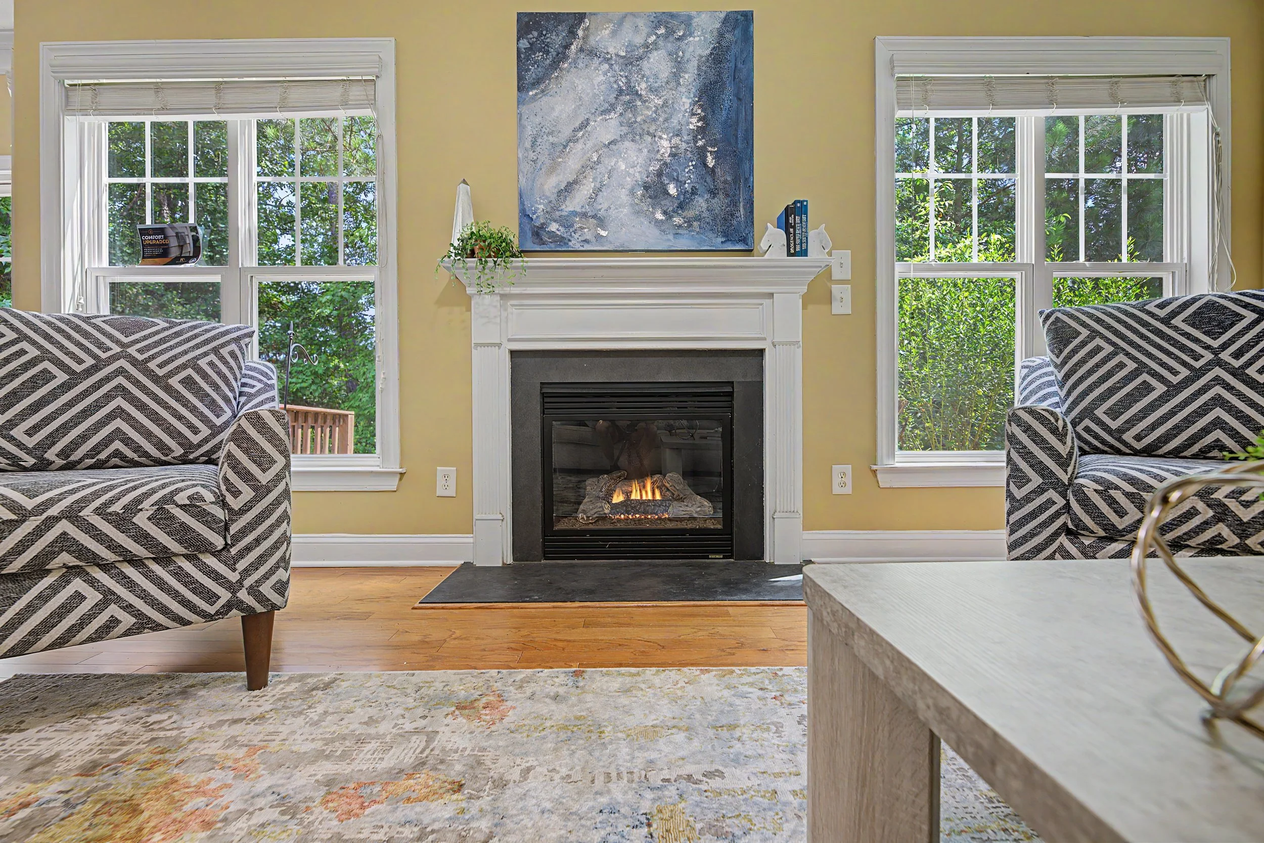 Living room with two armchairs featuring black and white geometric patterns, a fireplace with a fire burning, two large windows with white framing and blinds, a colorful abstract painting above the fireplace, a floating shelf with decorative items and books, a potted plant, and a section of a beige coffee table in the foreground.