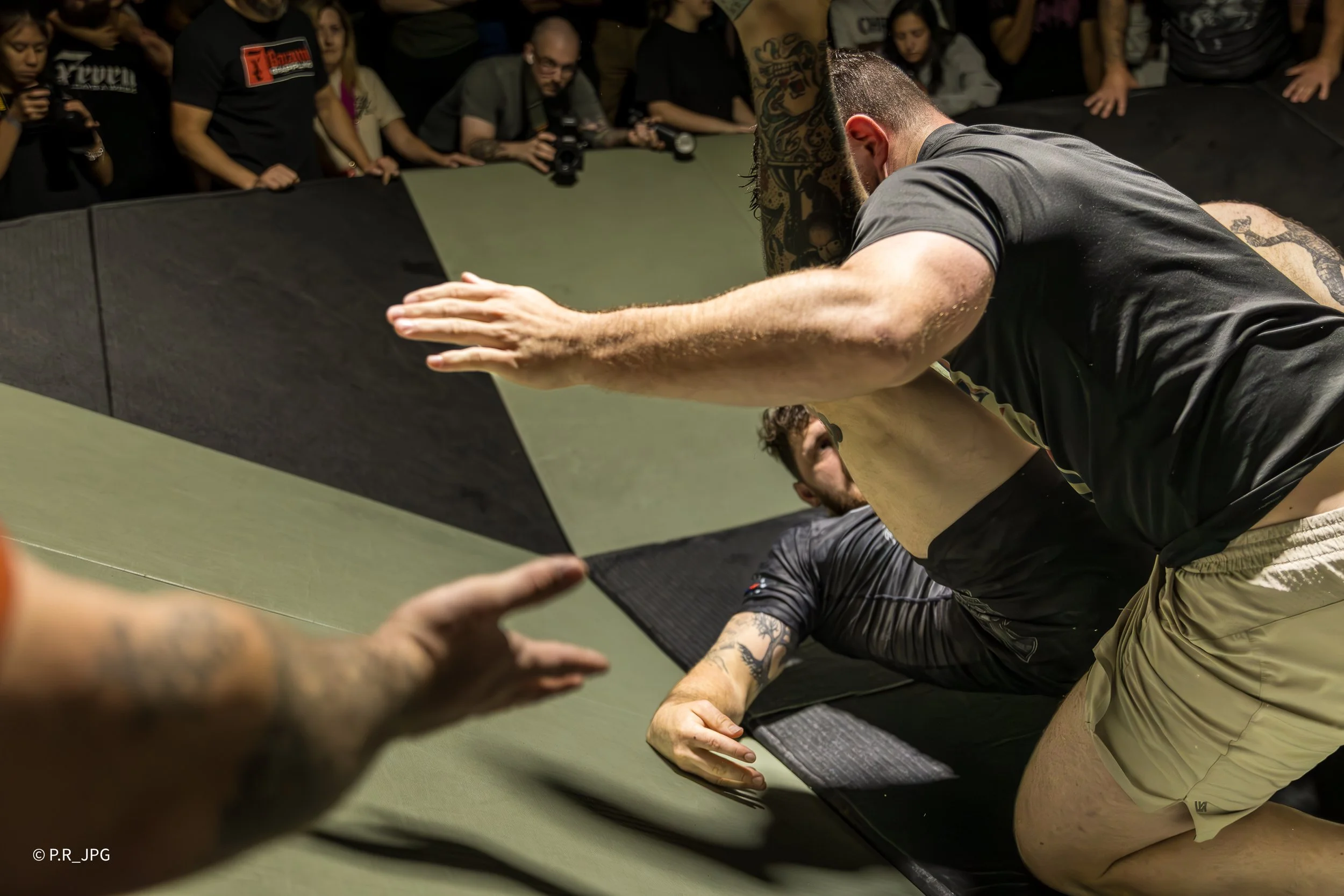 A group of people, some with tattoos, are gathered around a wrestling mat. Two men are engaged in a wrestling match; one is on top, pinning the other, both dressed in athletic clothing. Several spectators are watching closely, some taking photographs.