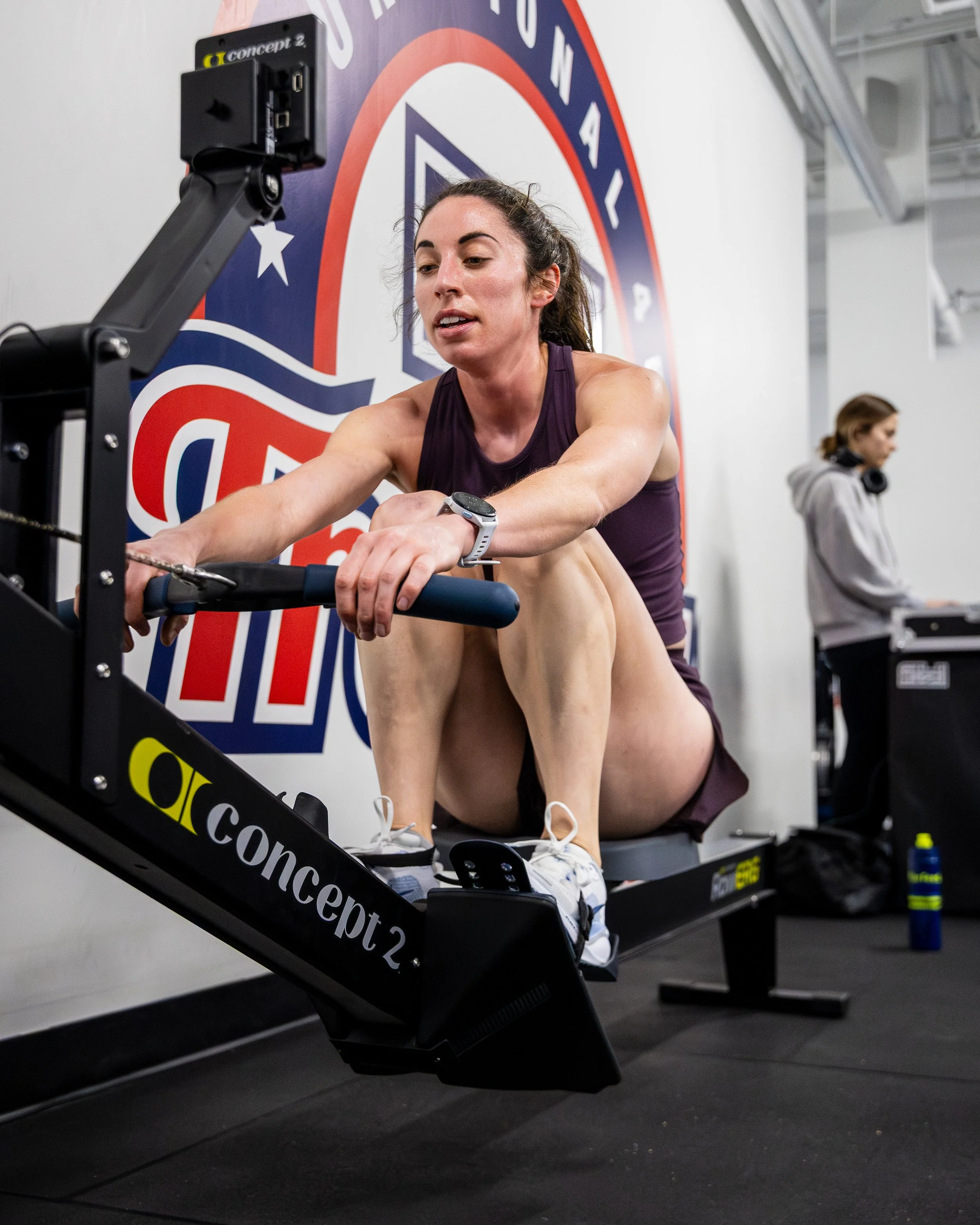 Woman exercising on a rowing machine in a gym, with a large logo on the wall behind her.