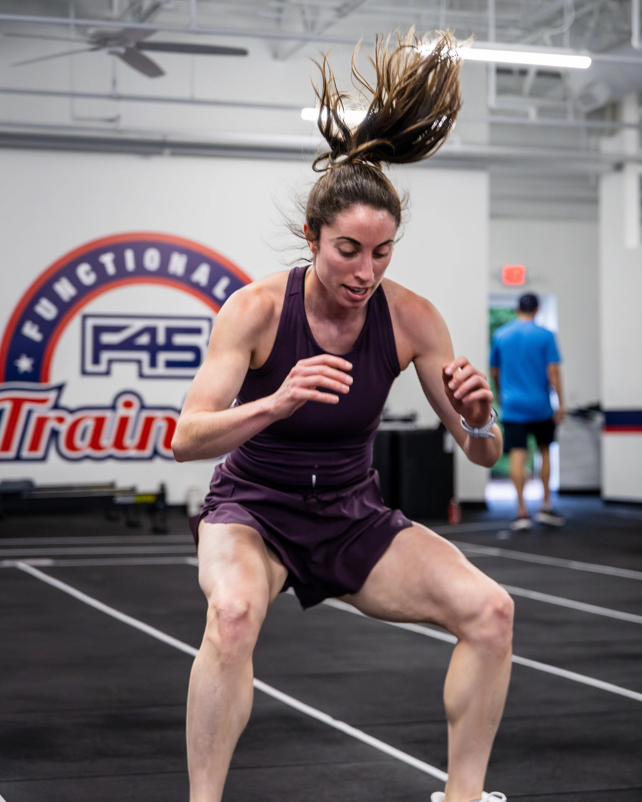 A woman with long hair in a ponytail wearing a purple tank top and shorts is exercising in a gym, with her hair flying upward and her face focused, with a blurry background of gym equipment and a person in a blue shirt.