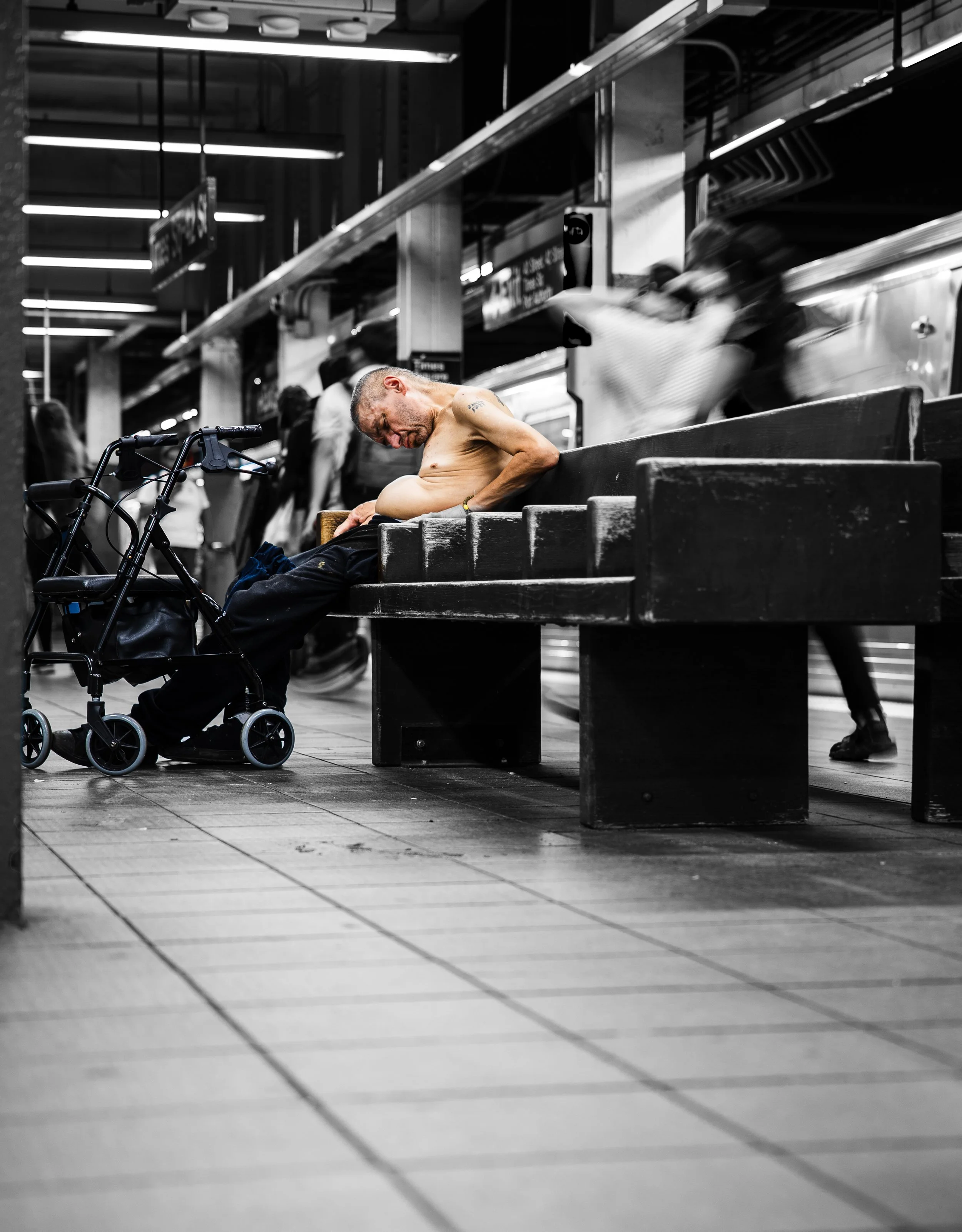 A man sitting shirtless on a bench in a subway station, appearing exhausted or unwell. A wheelchair and blurred individuals are nearby.