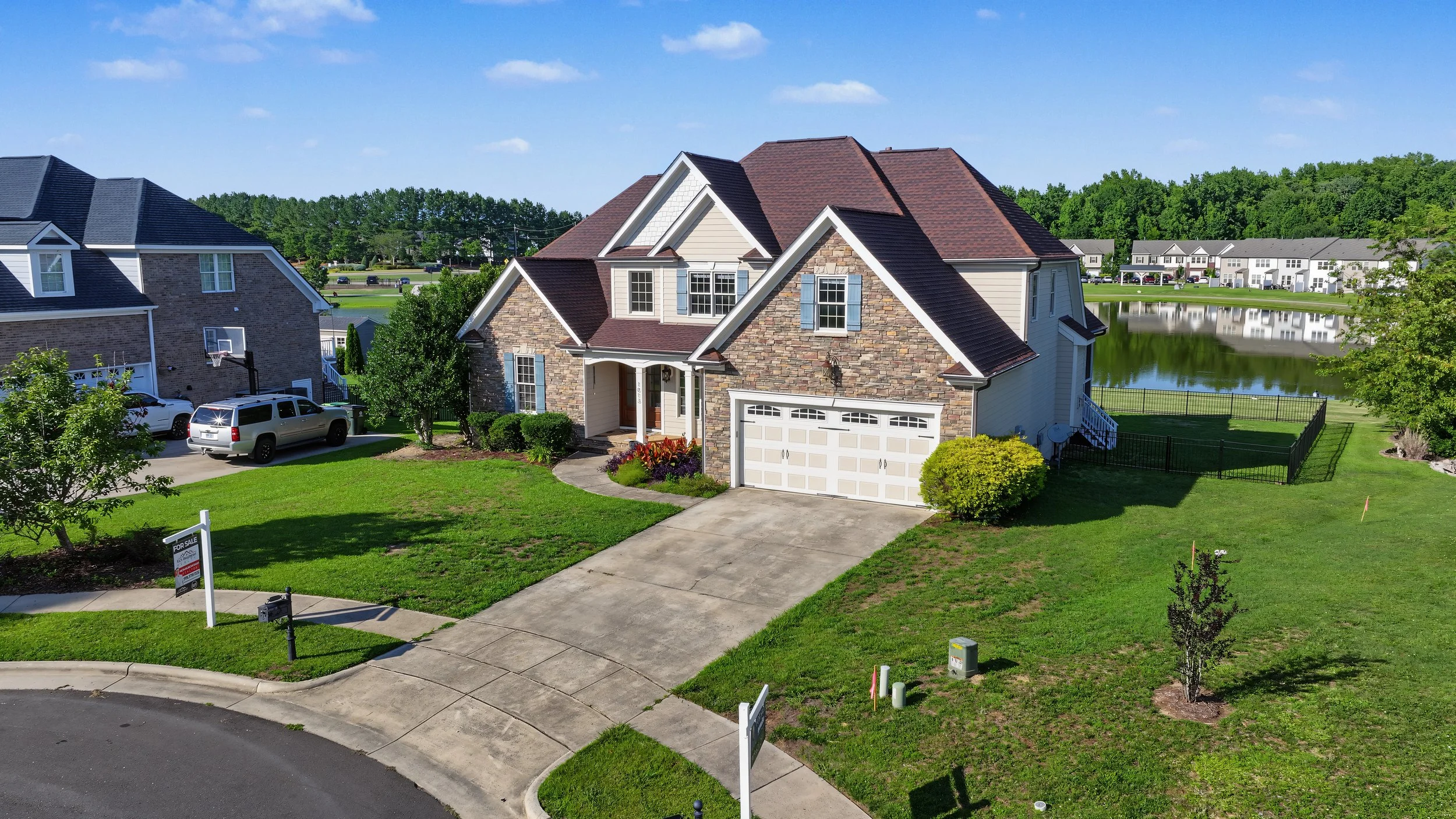A two-story house with a brick and siding exterior, attached garage, well-maintained lawn, and surrounding neighborhood with similar houses near a pond.