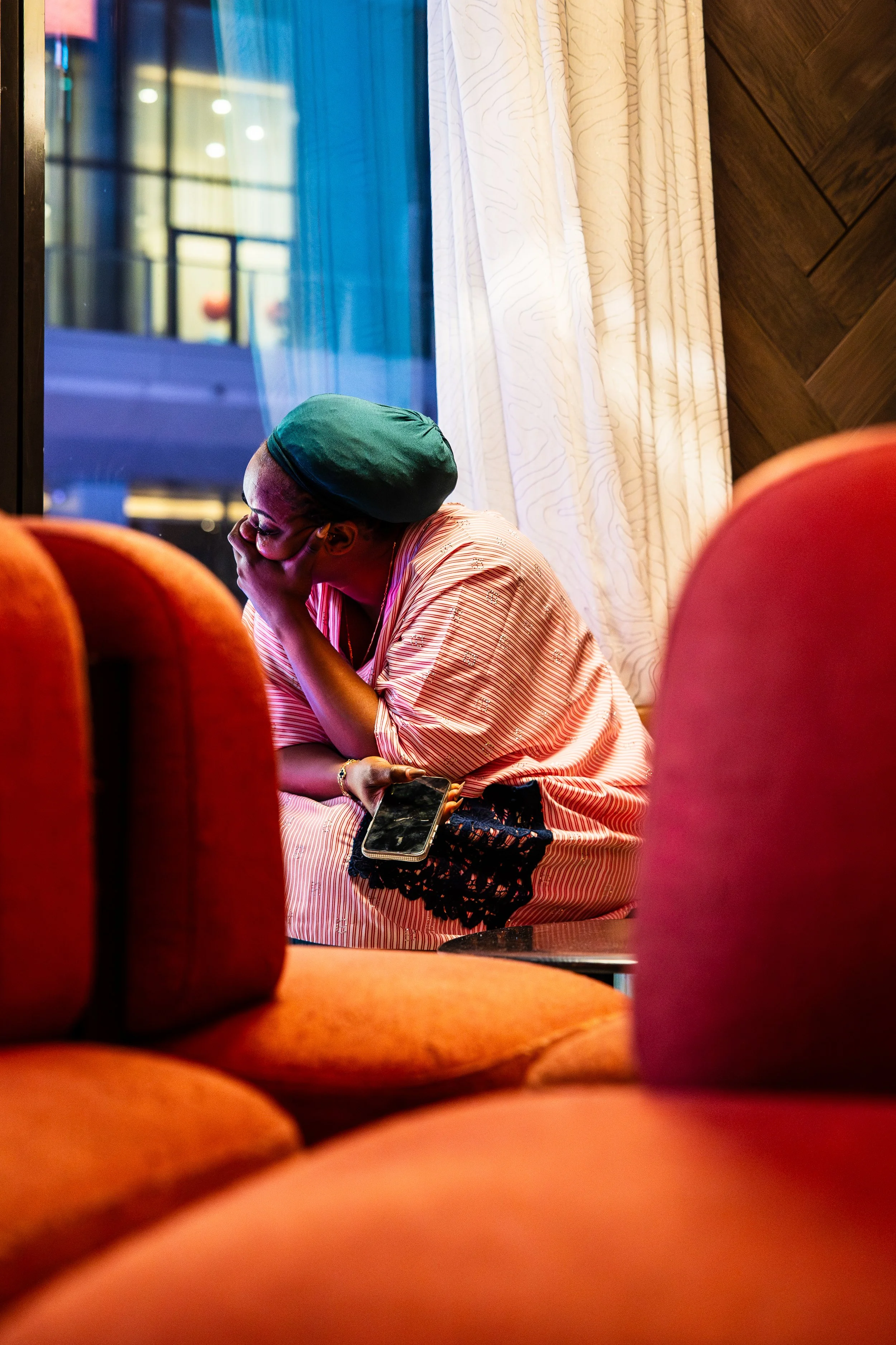 A woman with glasses and a dark green head covering, sitting by a window, resting her chin on her hand, holding a smartphone, with orange and pink chairs in the foreground and cream curtains and wooden wall in the background.