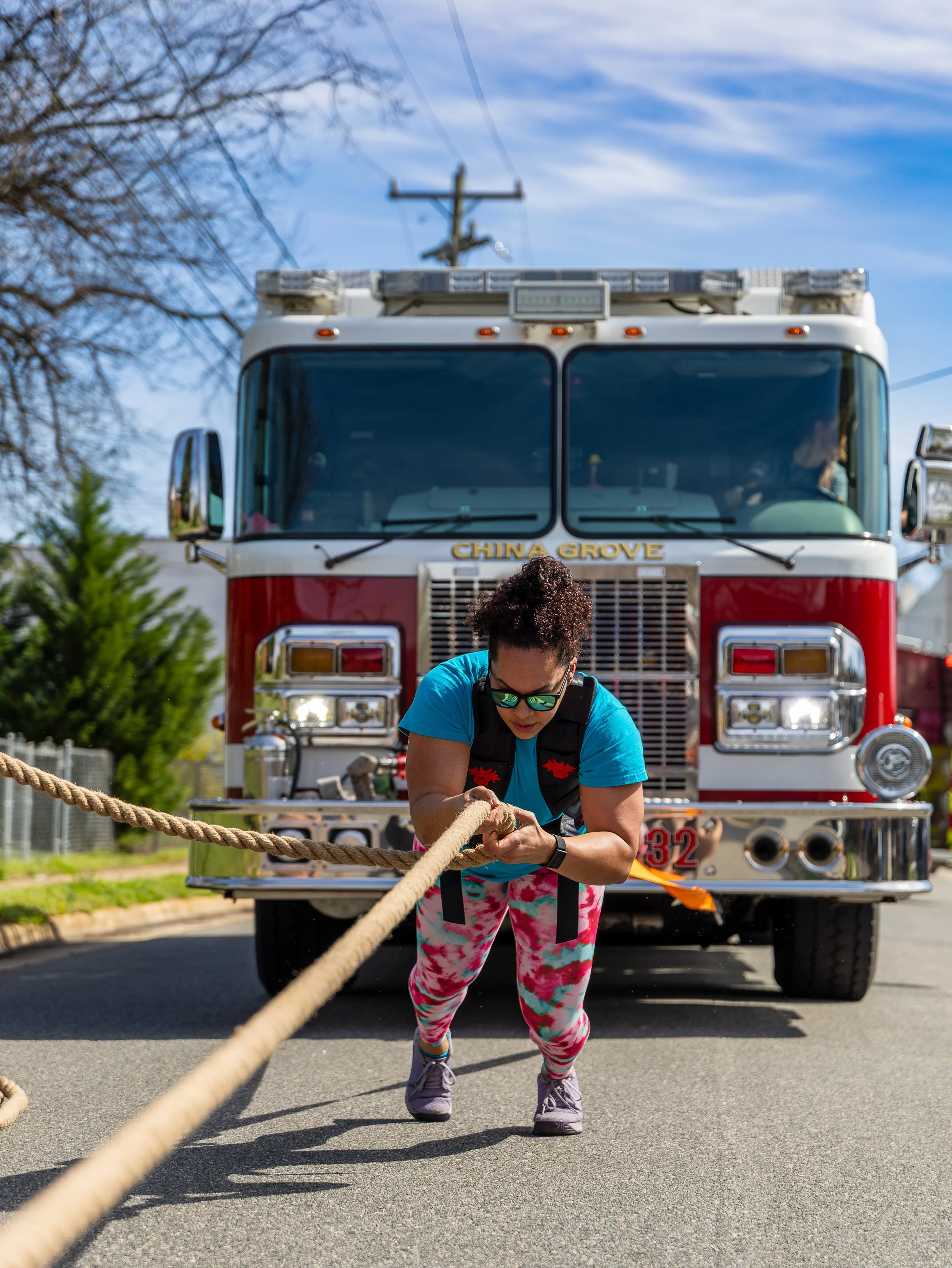A woman in colorful leggings, a blue shirt, and sunglasses is pulling a thick rope during a workout in front of a fire truck with the words 'China Grove' on it. The scene is outdoors with blue sky and trees.