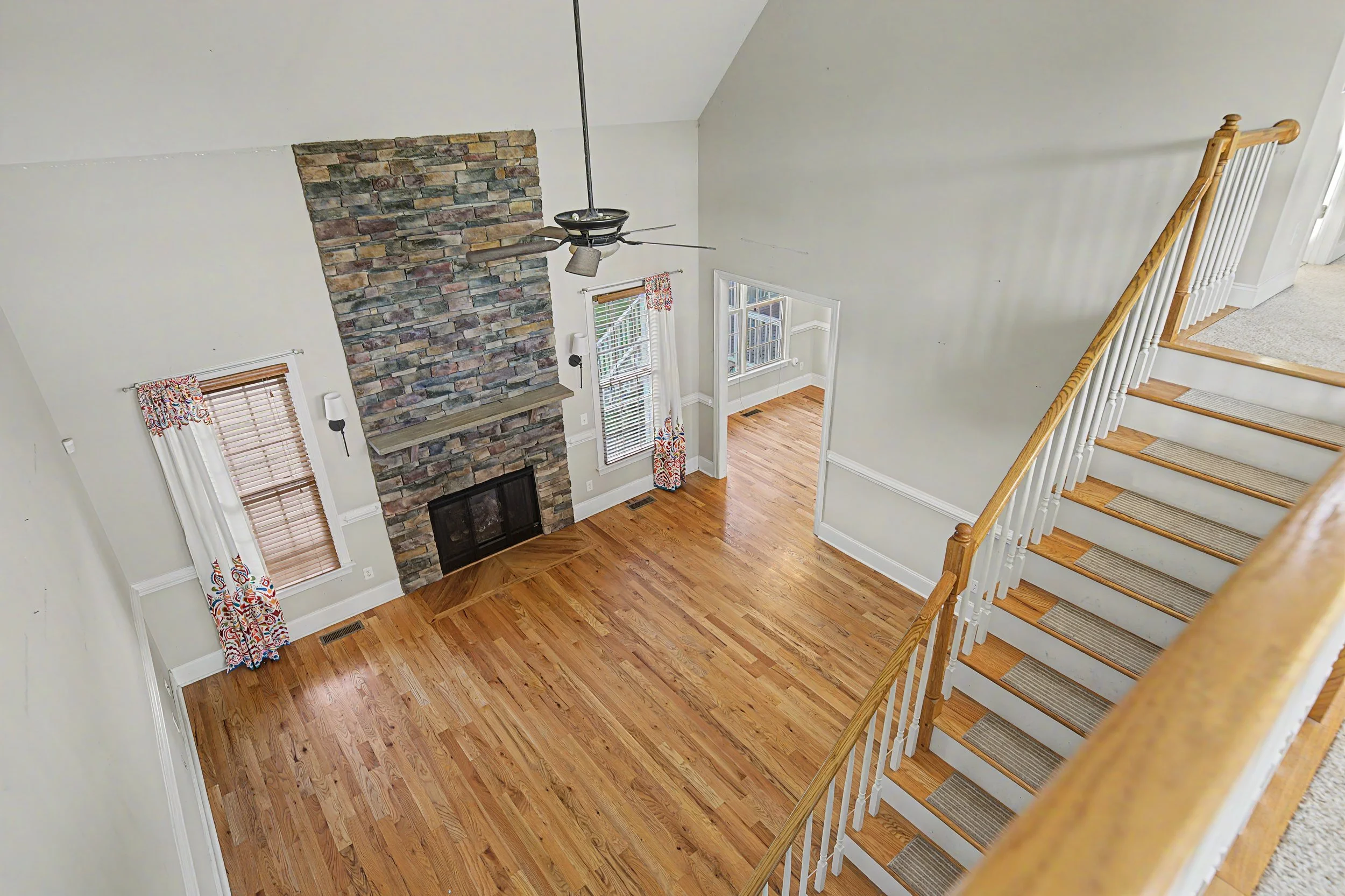 Interior view of a living room with a stone fireplace, wooden flooring, large windows with curtains, and a staircase leading upstairs.