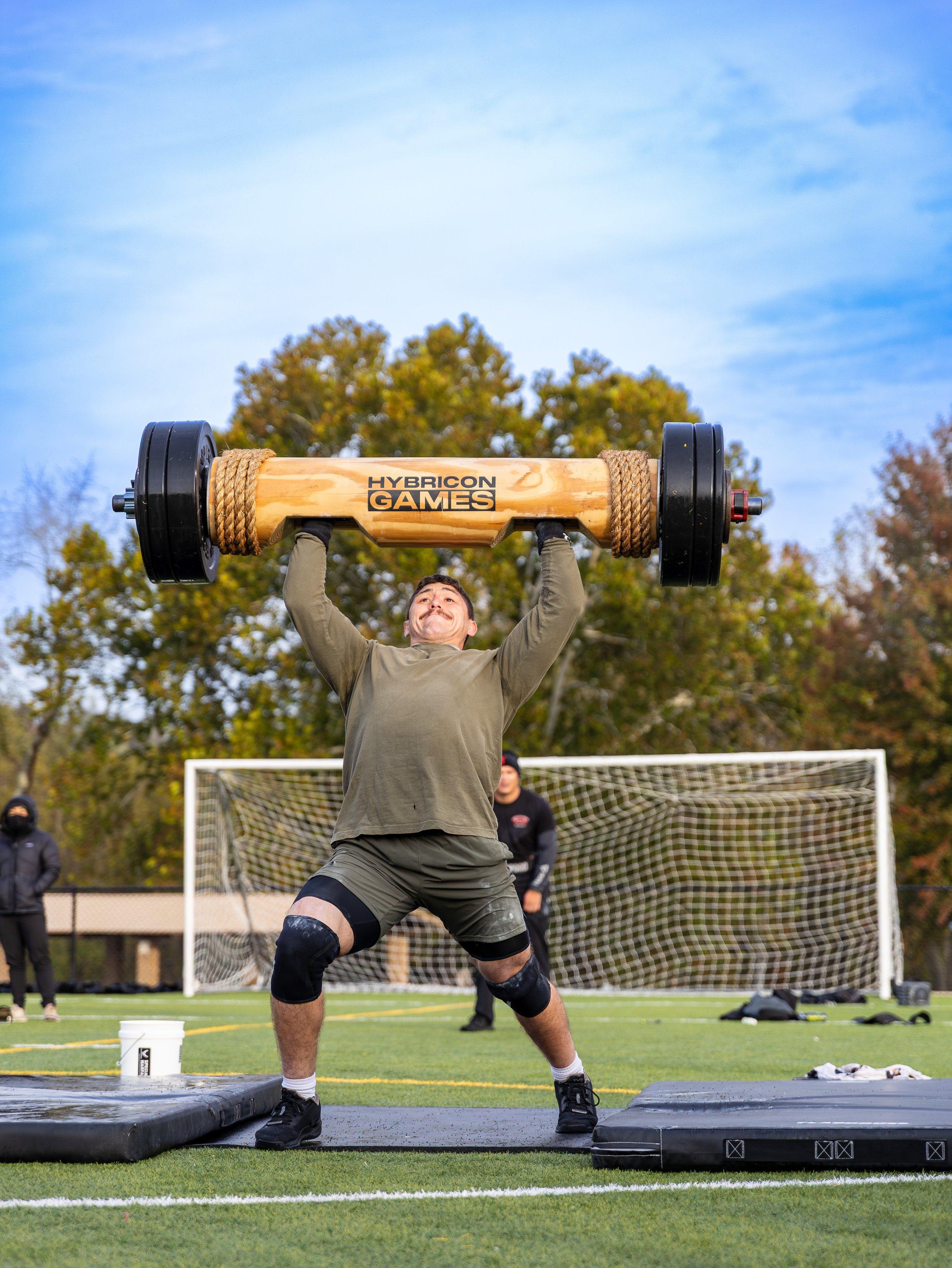 A man participating in an outdoor strength competition, lifting a large wooden log with the words "HYBRICON GAMES" on it while on a raised platform on a football field with a goalpost and trees in the background.