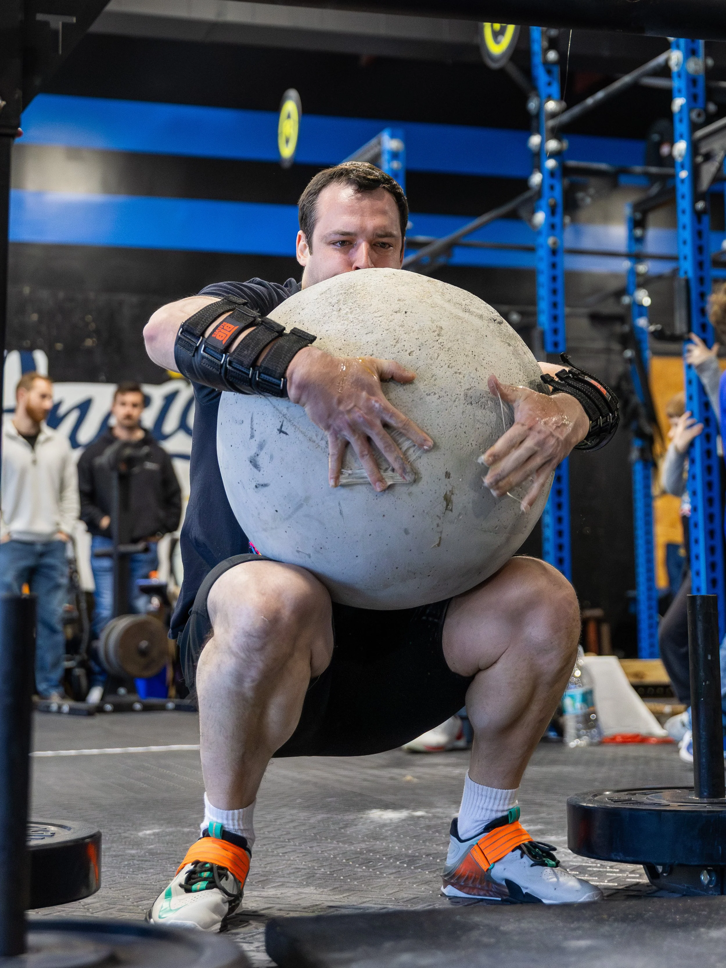 A man is performing a squat with a large stone in a gym. He is squatting in front of a squat rack, wearing athletic shoes, shorts, and a wrist brace. There are people and gym equipment in the background.