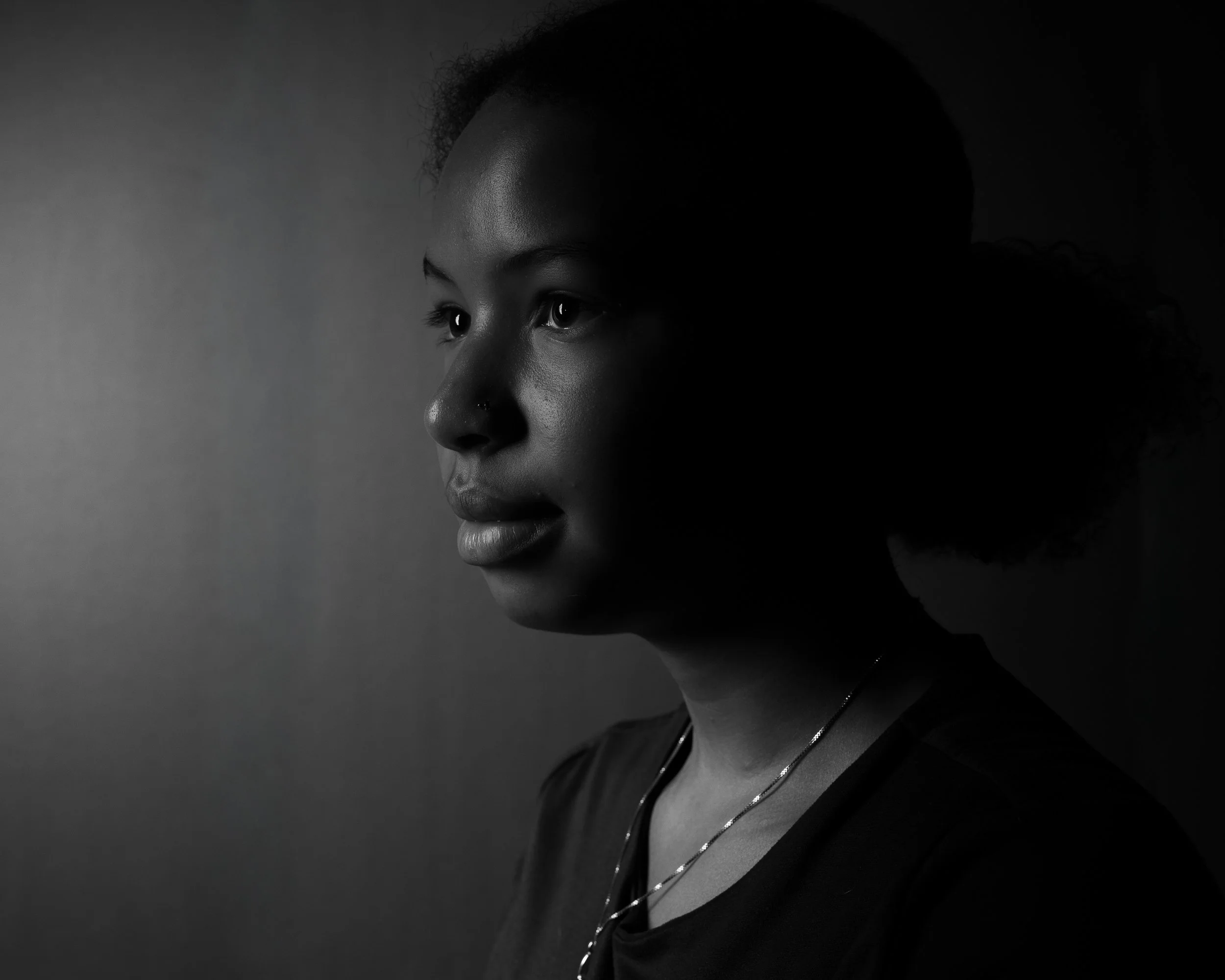 A black and white portrait of a woman with dark skin, curly hair, and wearing layered necklaces, looking thoughtfully to the side.