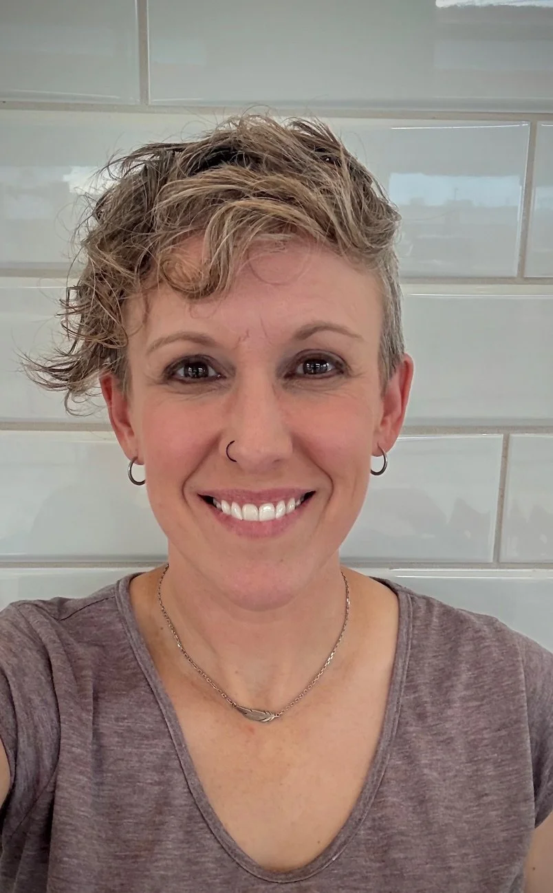 A smiling woman with short, curly, styled hair, wearing earrings, a nose ring, a necklace, and a T-shirt in front of a tiled wall.