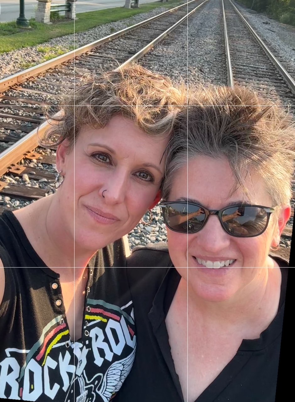 Dana and Kim taking a selfie in front of train tracks during sunset, smiling, one wearing sunglasses and the other with curly hair and a nose piercing.