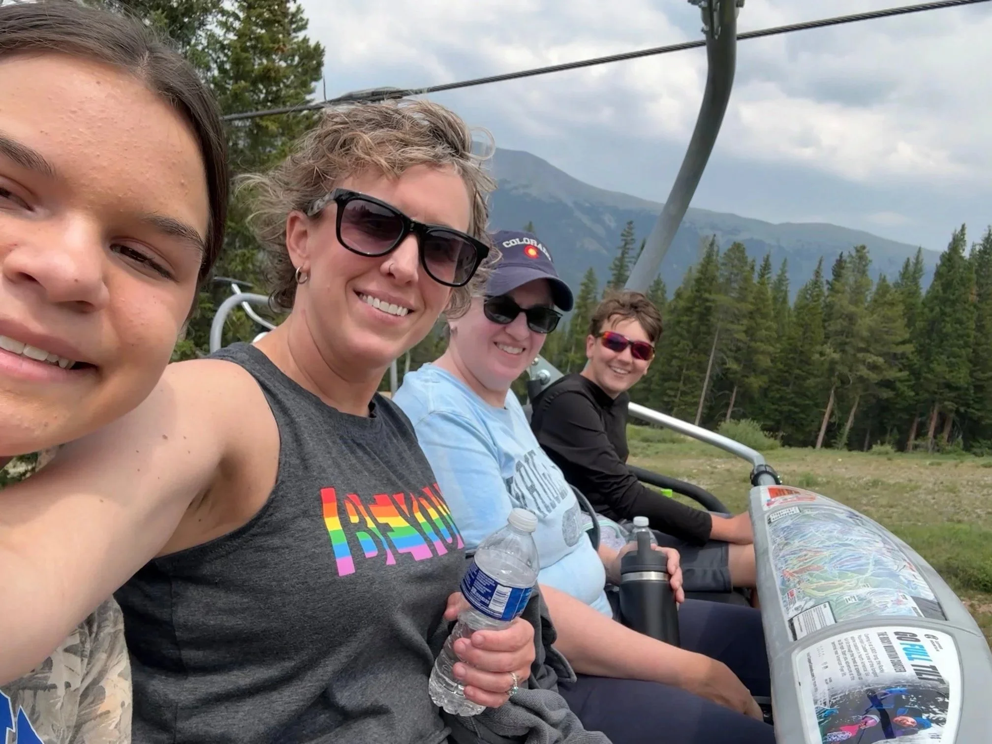 Four people sitting on a ski lift, smiling and holding water bottles, with a mountain and trees in the background.