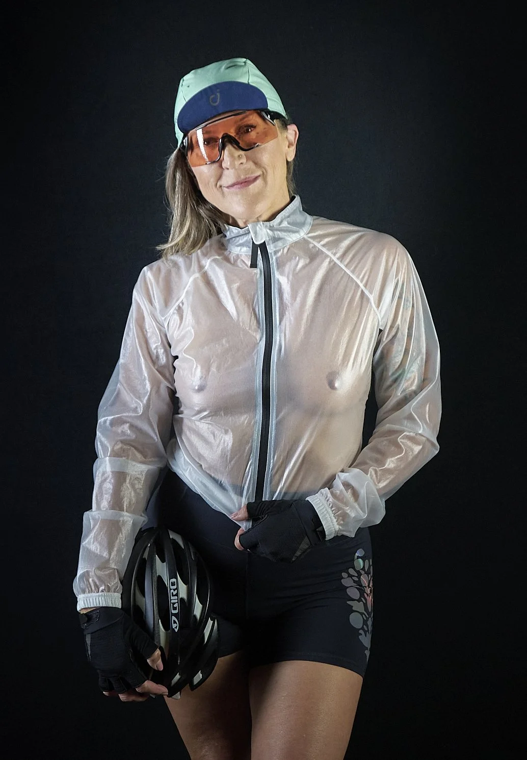 A woman dressed in athletic biking gear holding a helmet, wearing a white windbreaker, black gloves, black shorts with a pattern, and a cycling cap with glasses, standing against a plain dark background.